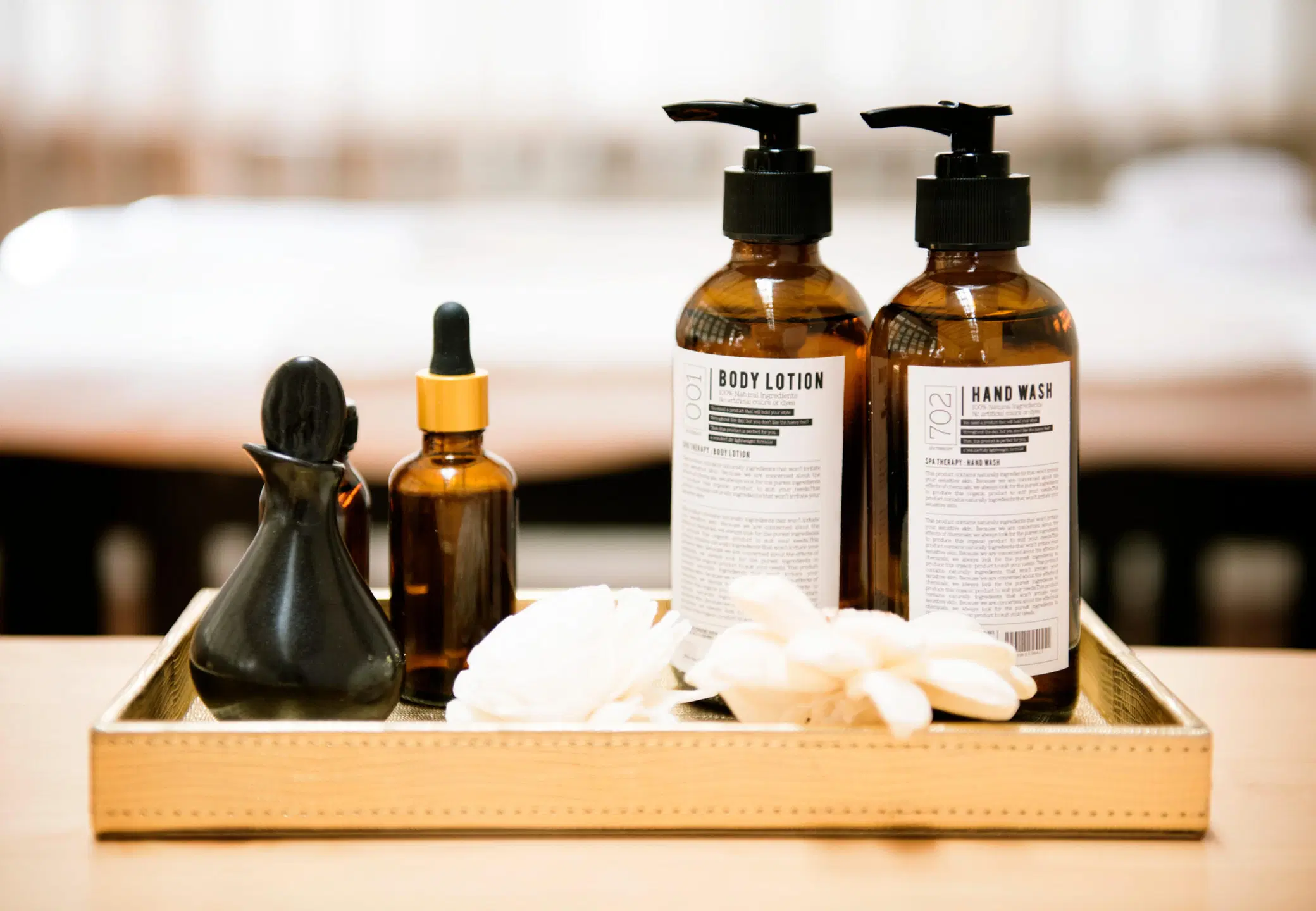 Kultaisella tarjottimella ruskeat pumppupullot, joissa lukee Body Lotion ja Hand Wash, sekä pipettipullo ja vaaleita kukkia.