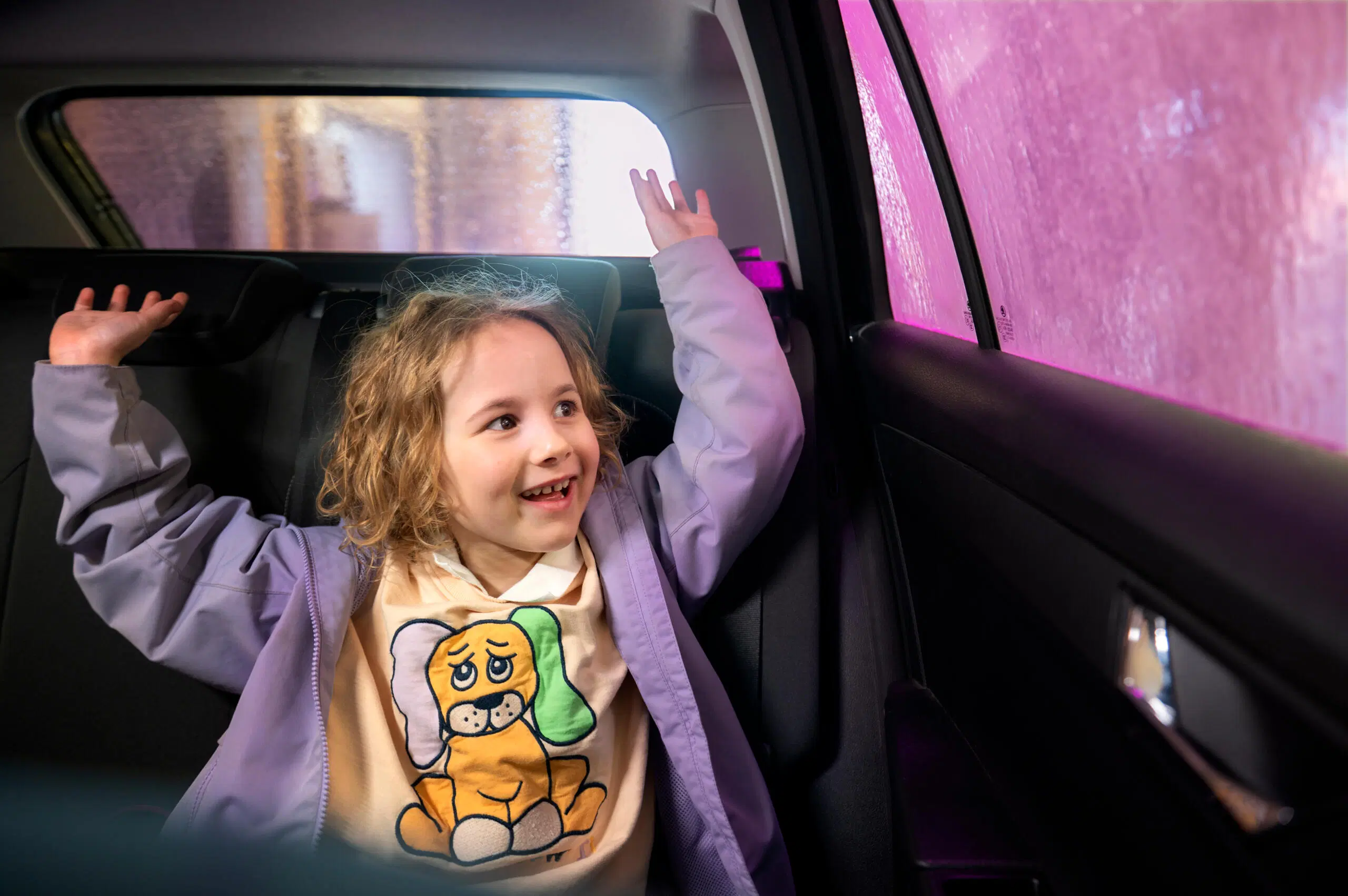 Iloinen lapsi nostaa käsiään autopesussa auton takapenkillä, pesutunneli ikkunan takana.