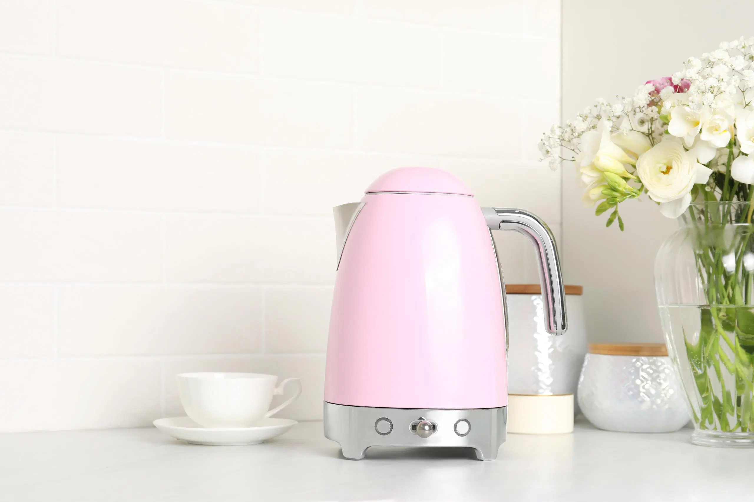 Vaaleanpunainen vedenkeitin, valkoinen kahvikuppi, kaksi purkkia ja kukkia maljakossa keittiötasolla.