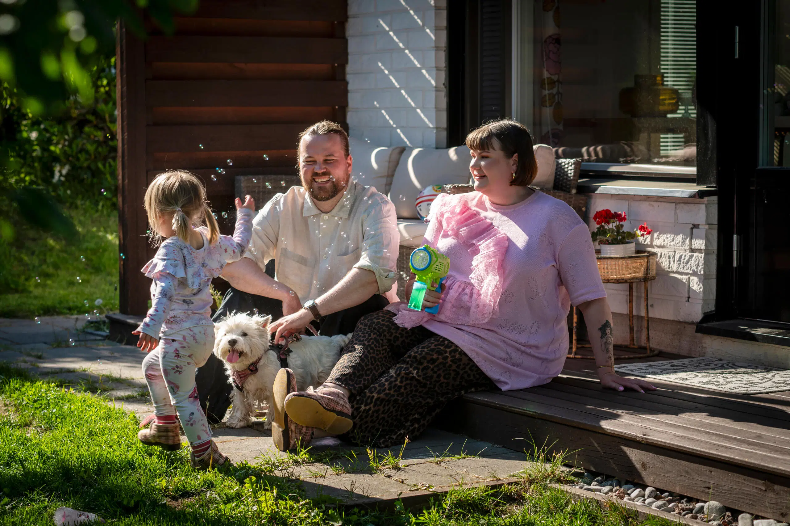 Perhe istuu portailla auringossa, lapsi puhaltaa saippuakuplia, pieni valkoinen koira mukana pihalla.