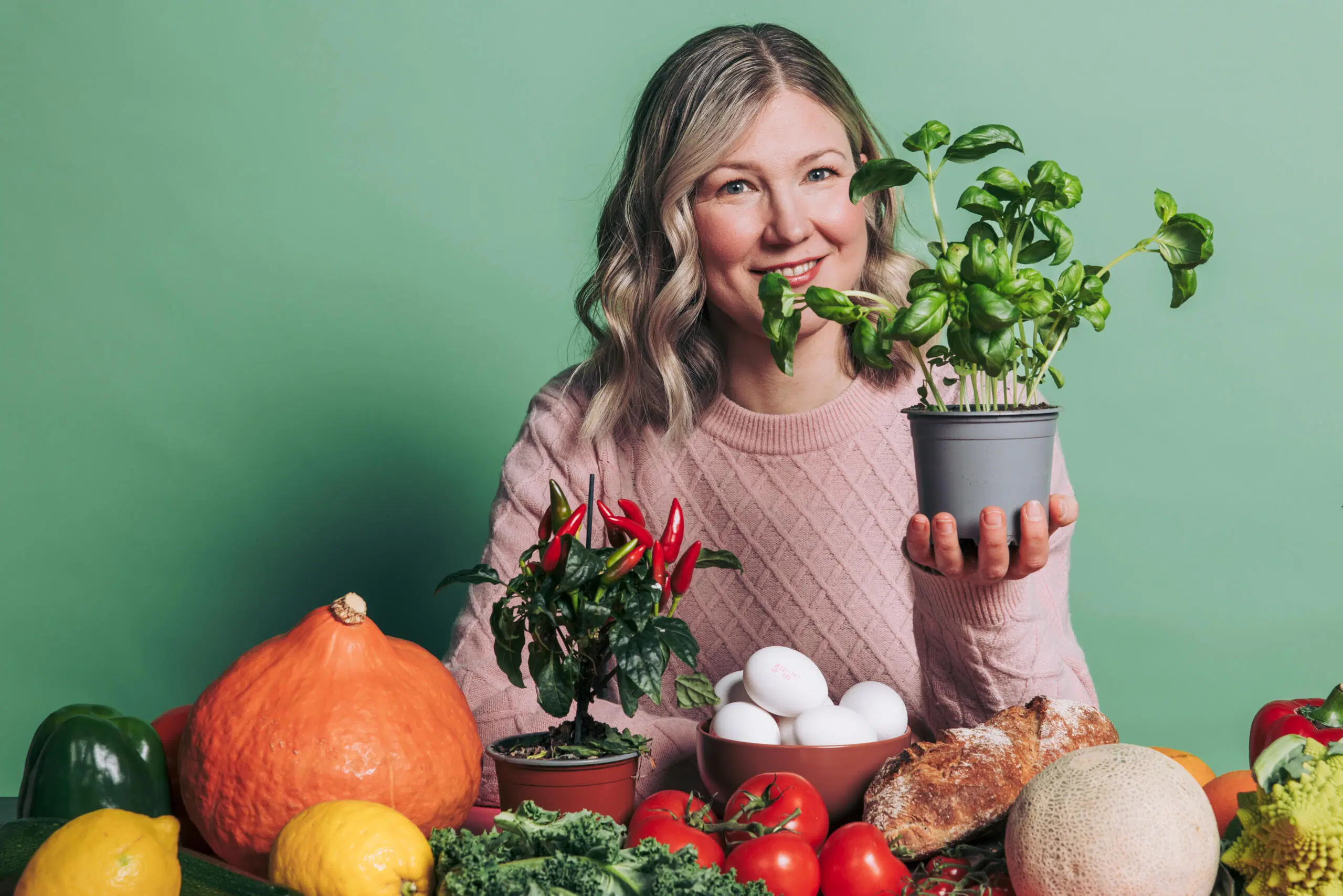 Hymyilevä henkilö pitää basilikaa ruukussa värikkäiden vihannesten ja hedelmien keskellä vihreää taustaa vasten.
