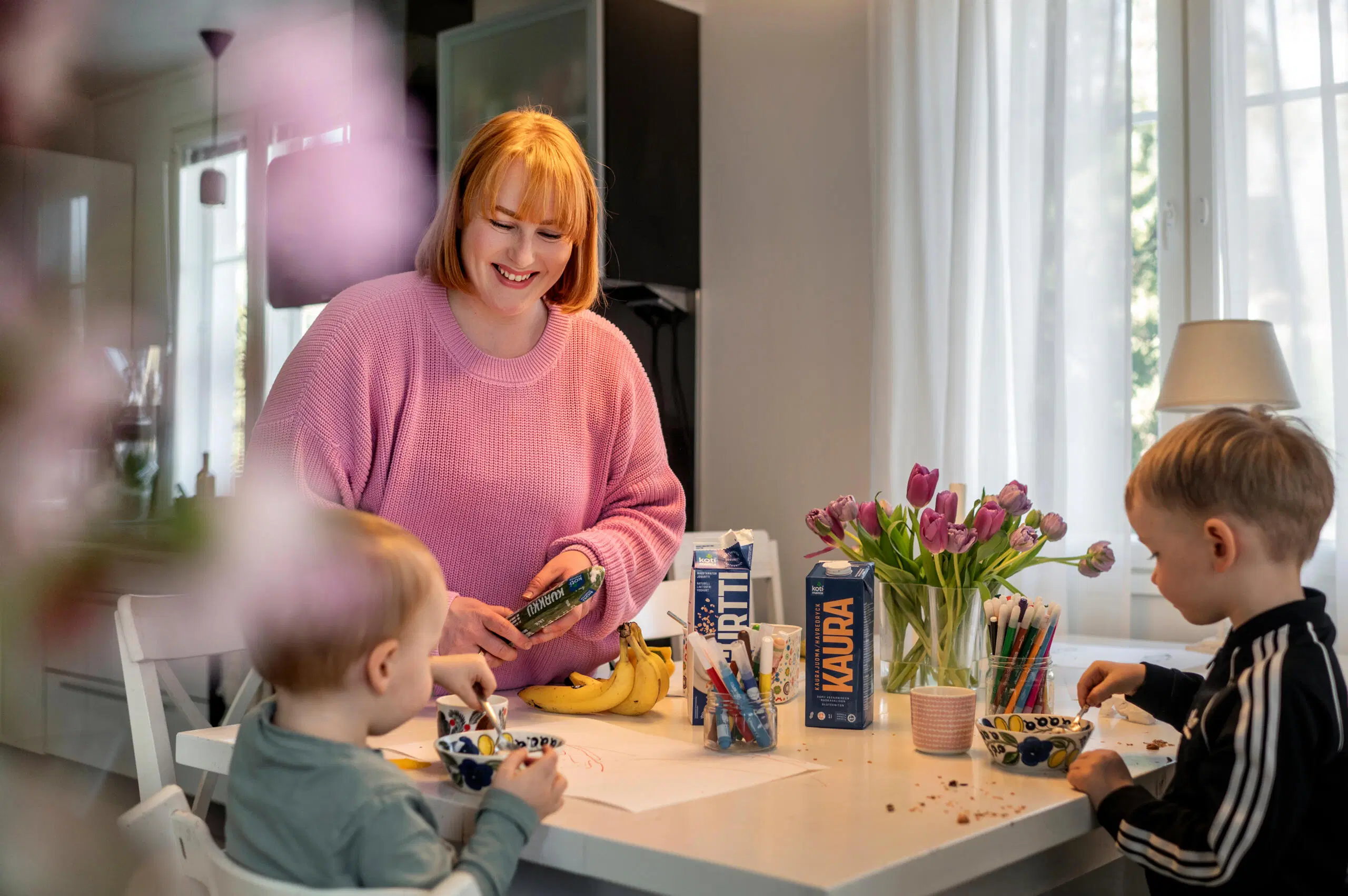 Nainen hymyilee keittiössä kahden lapsen kanssa pöydän ääressä; välipaloja, kukkia ja värikkäitä pakkauksia.