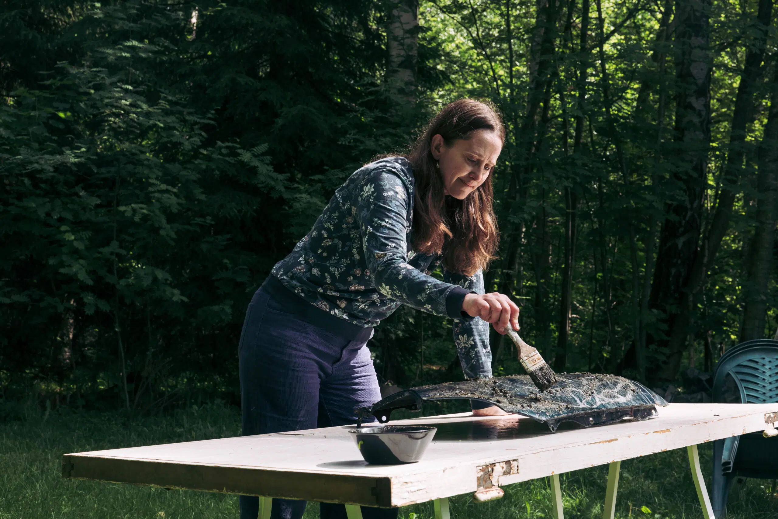 Sonja Salomäki maalaa mustalla maalilla ulkona pöydällä, metsä taustalla.