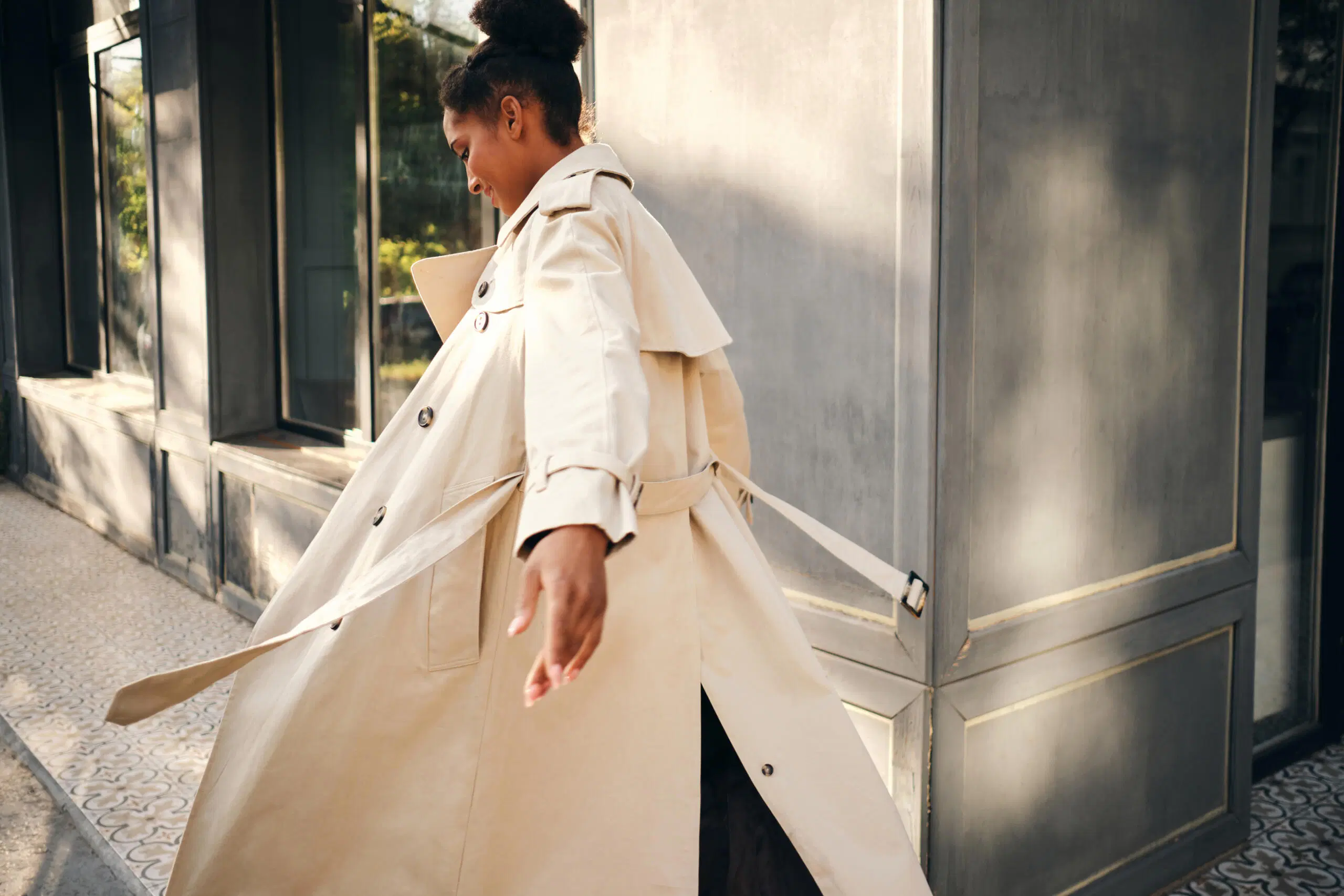 Nainen kävelee aurinkoisella kadulla vaalea trenssitakki hulmuten.