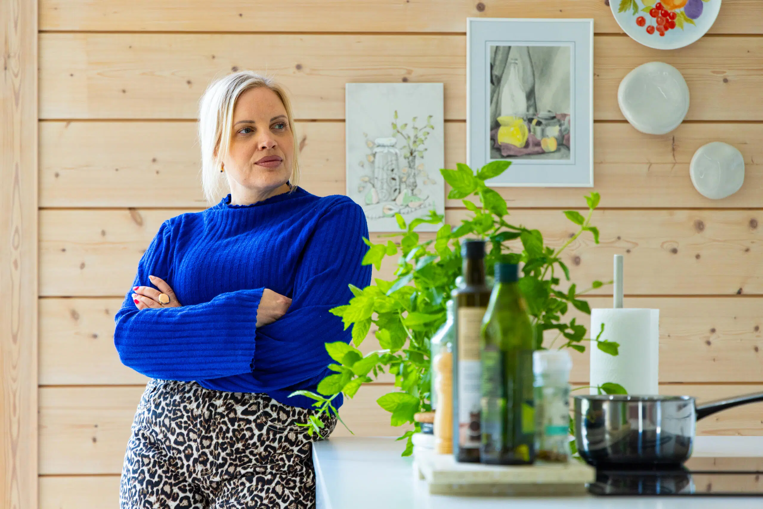 Nainen sinisessä neuleessa seisoo keittiössä käsivarret ristissä; edessä yrttejä ja oliiviöljypulloja puupaneeliseinää vasten.