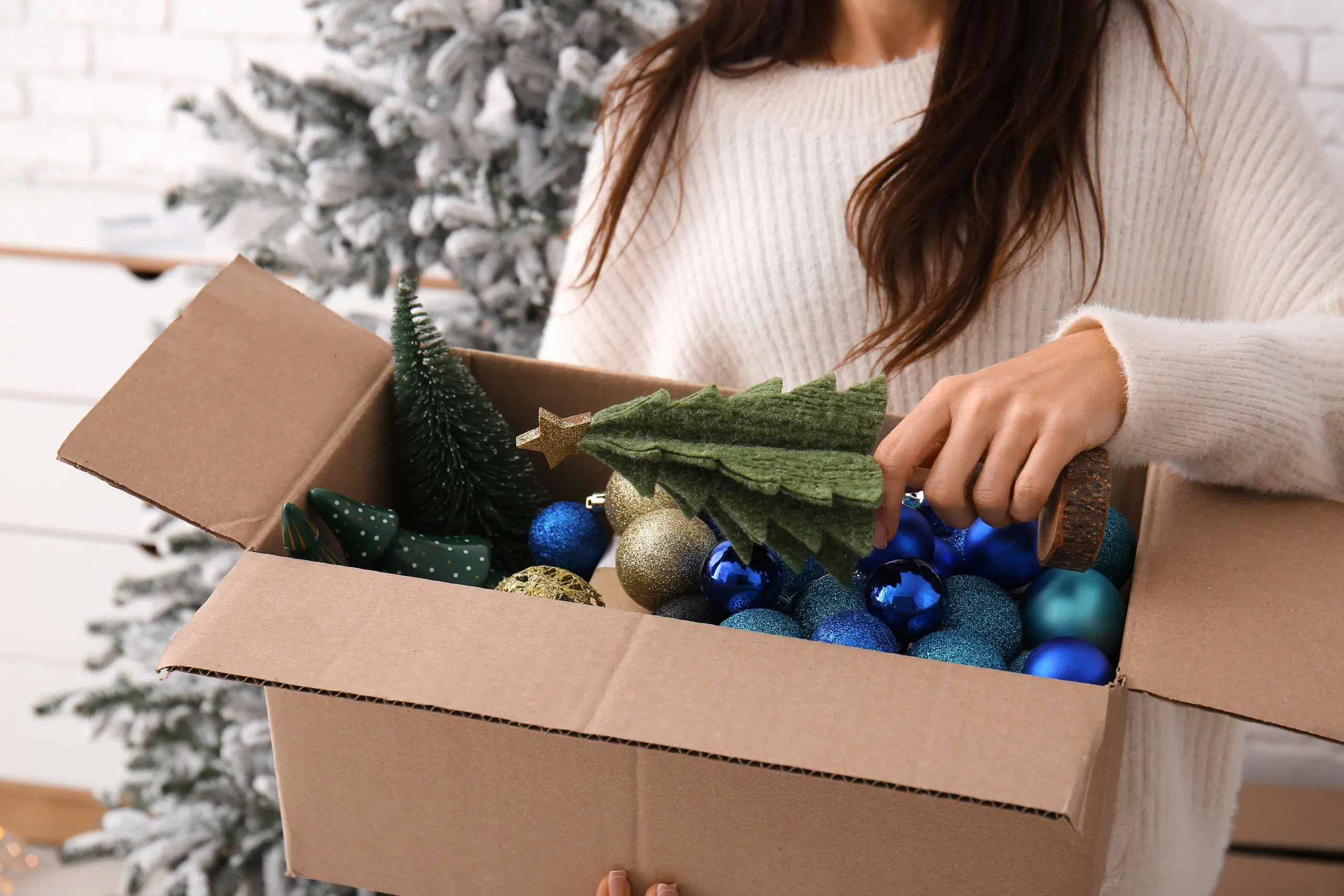 Naisen käsi laittaa koristekuusta pahvilaatikkoon, jossa on joulukoristeita