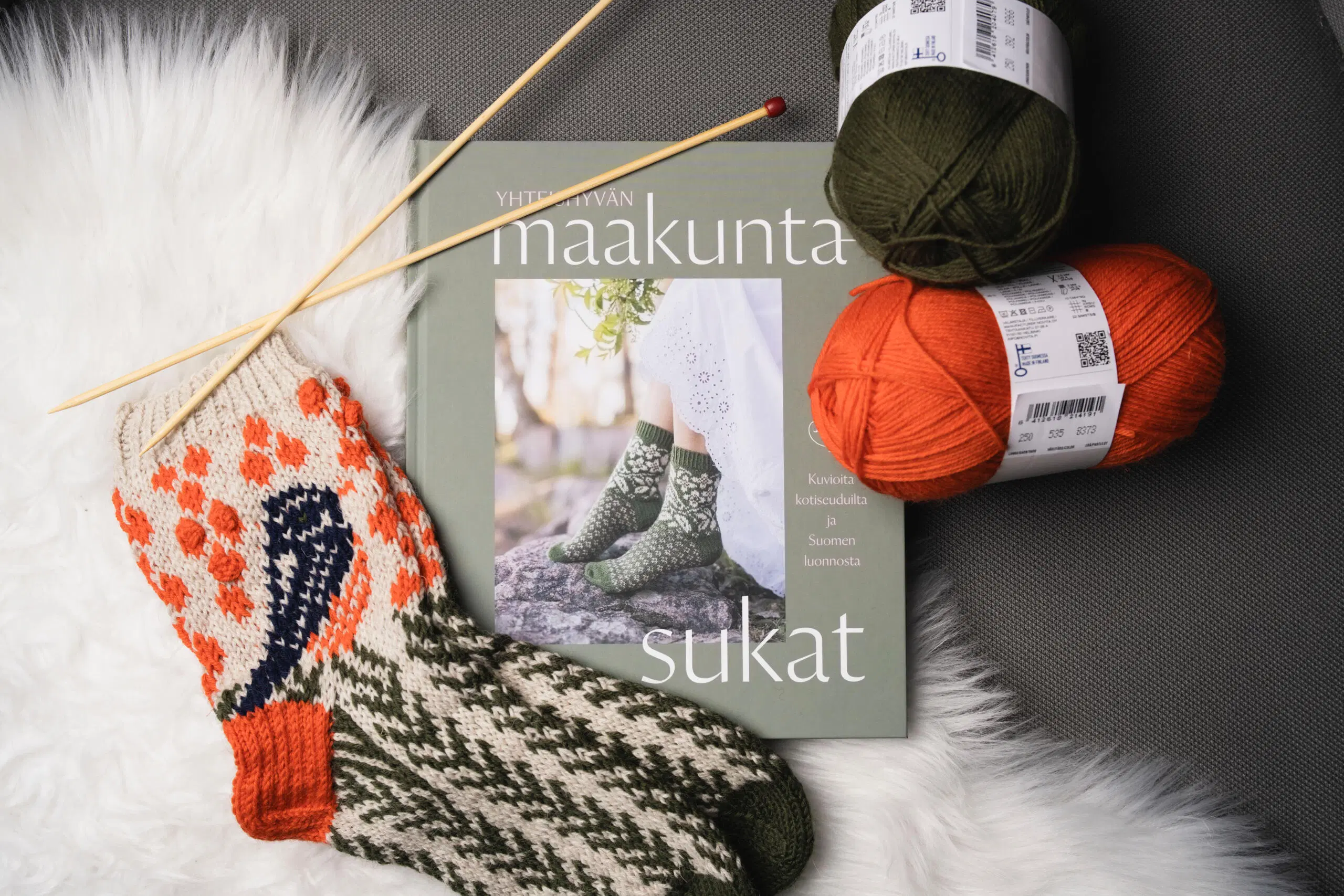 Yhteishyvän maakuntasukat -kirja, kaksi lankakerää, villasukat ja neulepuikot.