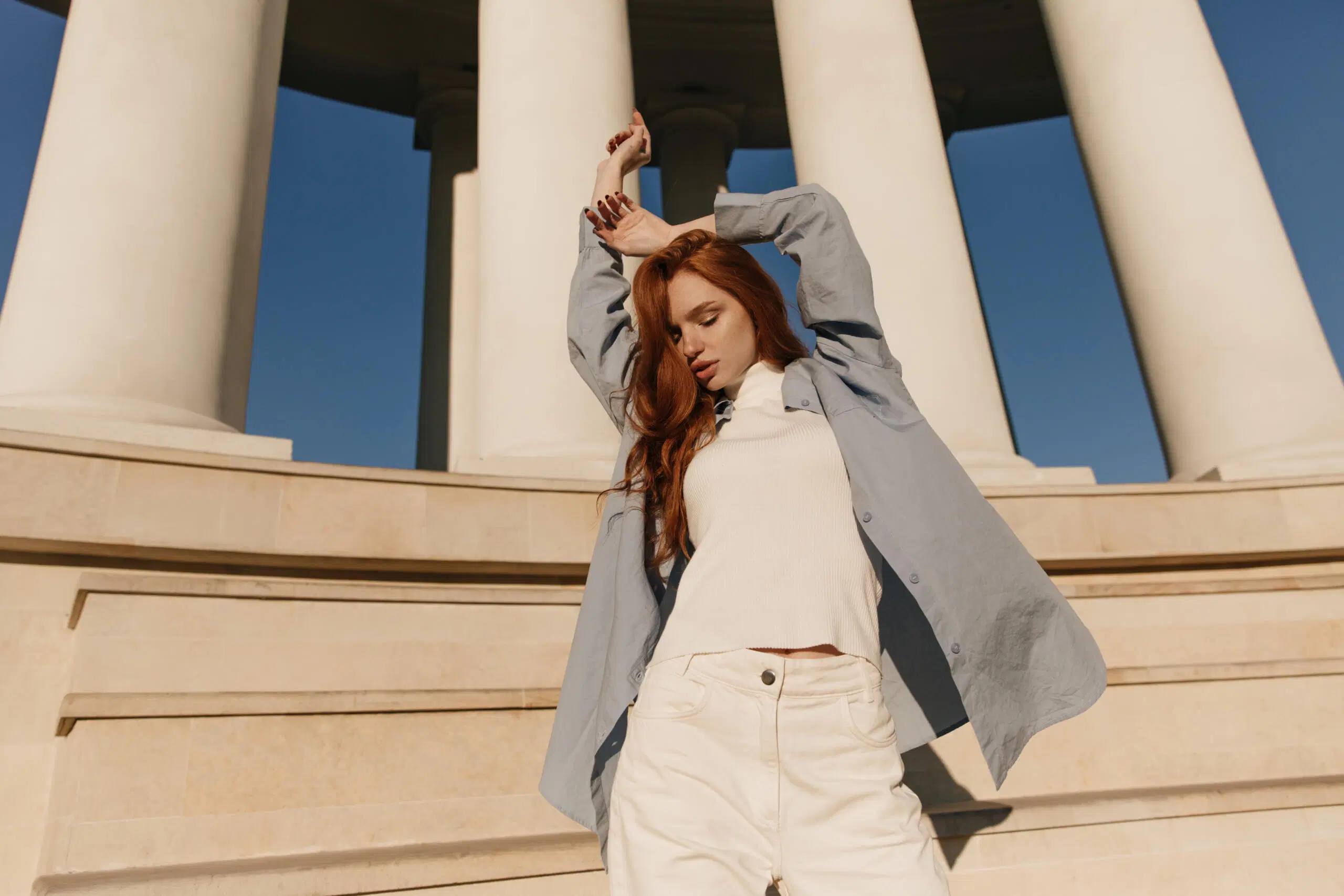 Nainen, jolla on asussaan vaaleansinistä ja valkoista. Poseeraa auringonpaisteessa kädet pään yläpuolella.