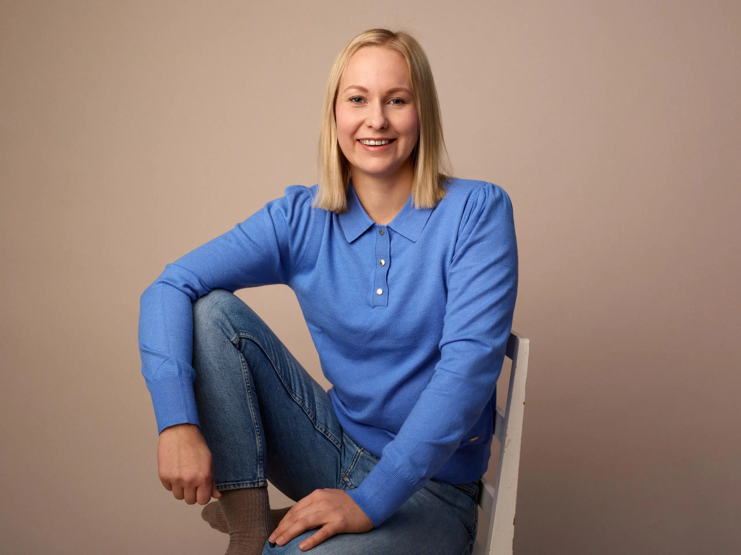 Elina Vessonen sinisessä neuleessa istuu tuolilla jalka ylhäällä, vaalea tausta.