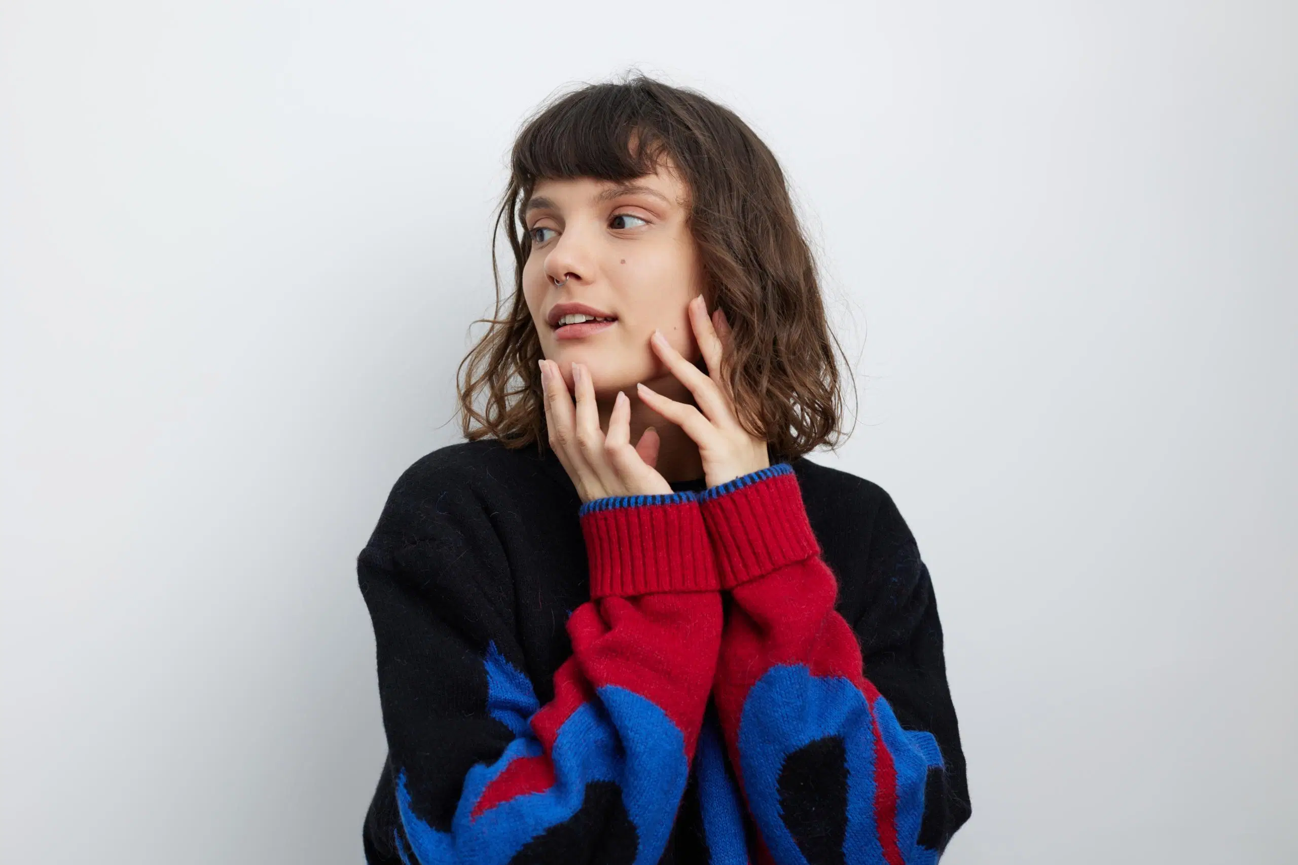 Värikkääseen neuleeseen pukeutunut nainen katsoo sivulle ja kehystää kasvoja käsillään