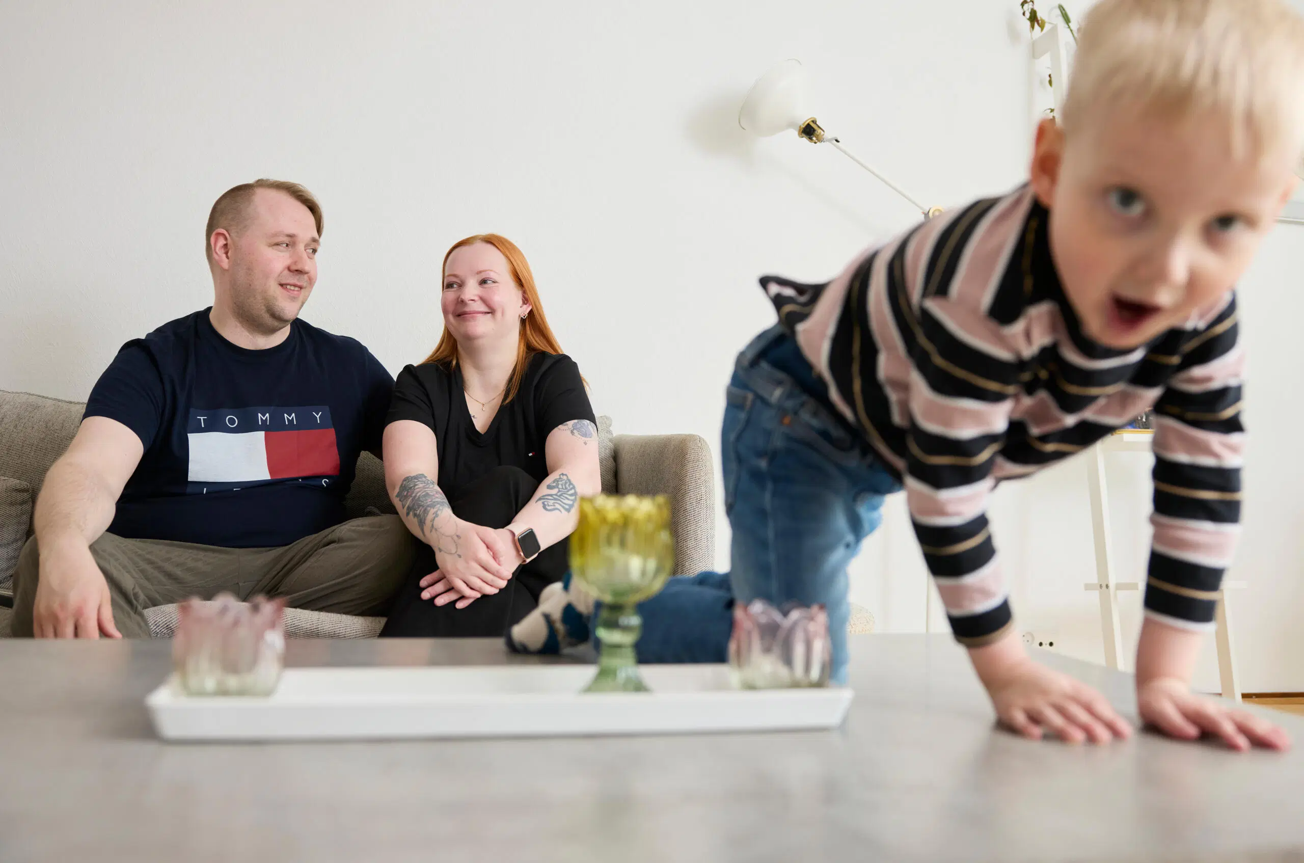 Pieni lapsi kiipeää pöydälle etualalla, vanhemmat istuvat sohvalla taustalla valoisassa olohuoneessa.