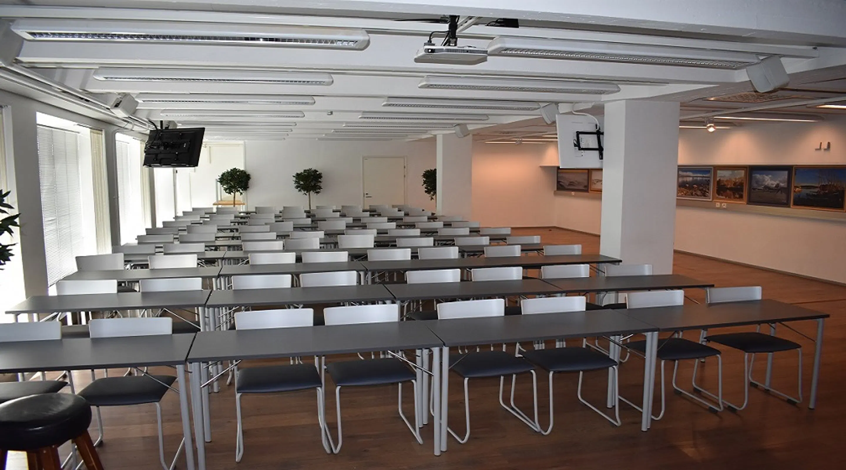 Bright meeting room with long tables and chairs arranged in rows. Wooden floor, small potted trees, and a display area with framed pictures on the wall. Ceiling projector installed.