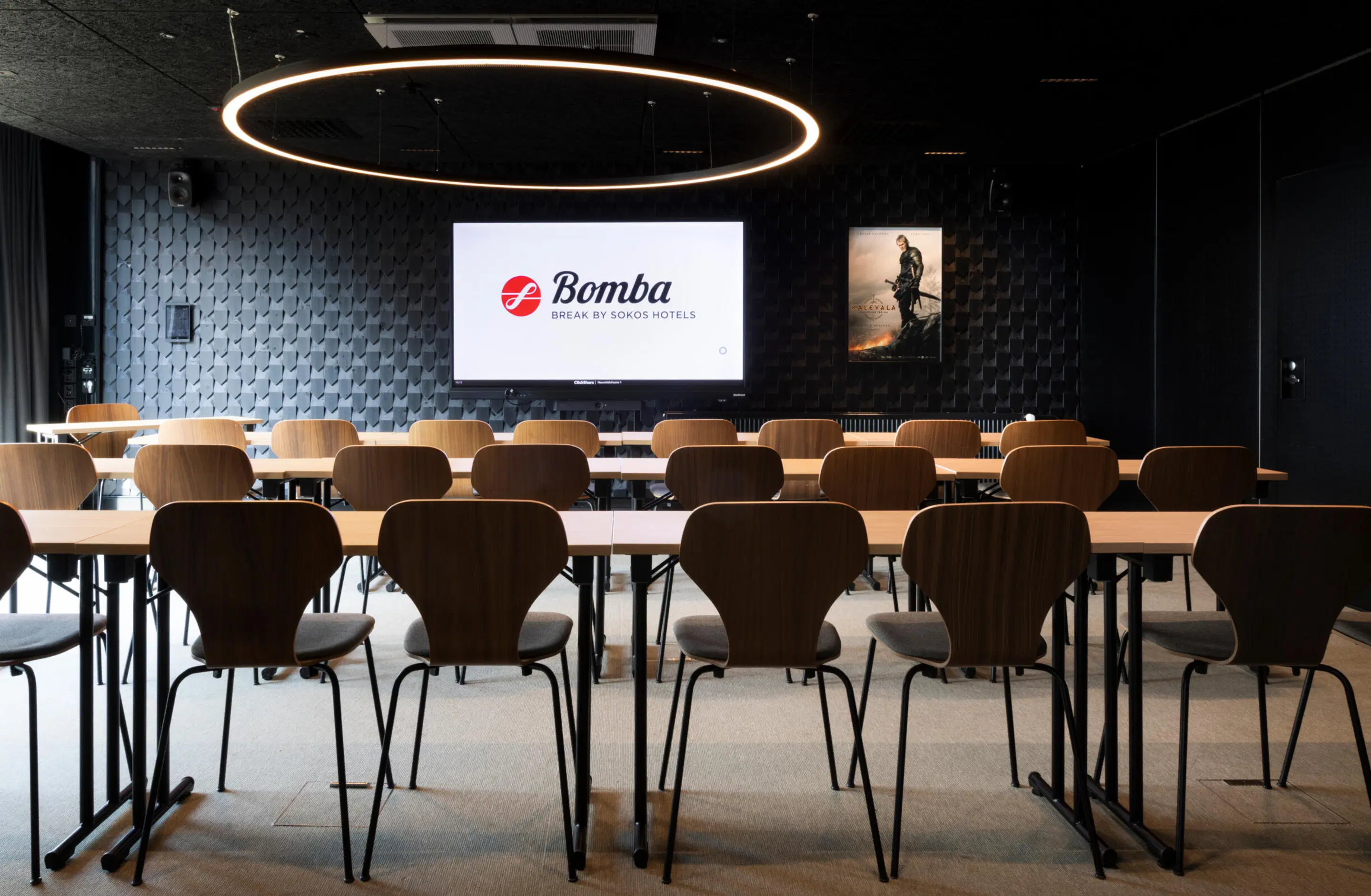 Modern conference room with long tables and wooden chairs, large screen, circular pendant light and dark textured wall.