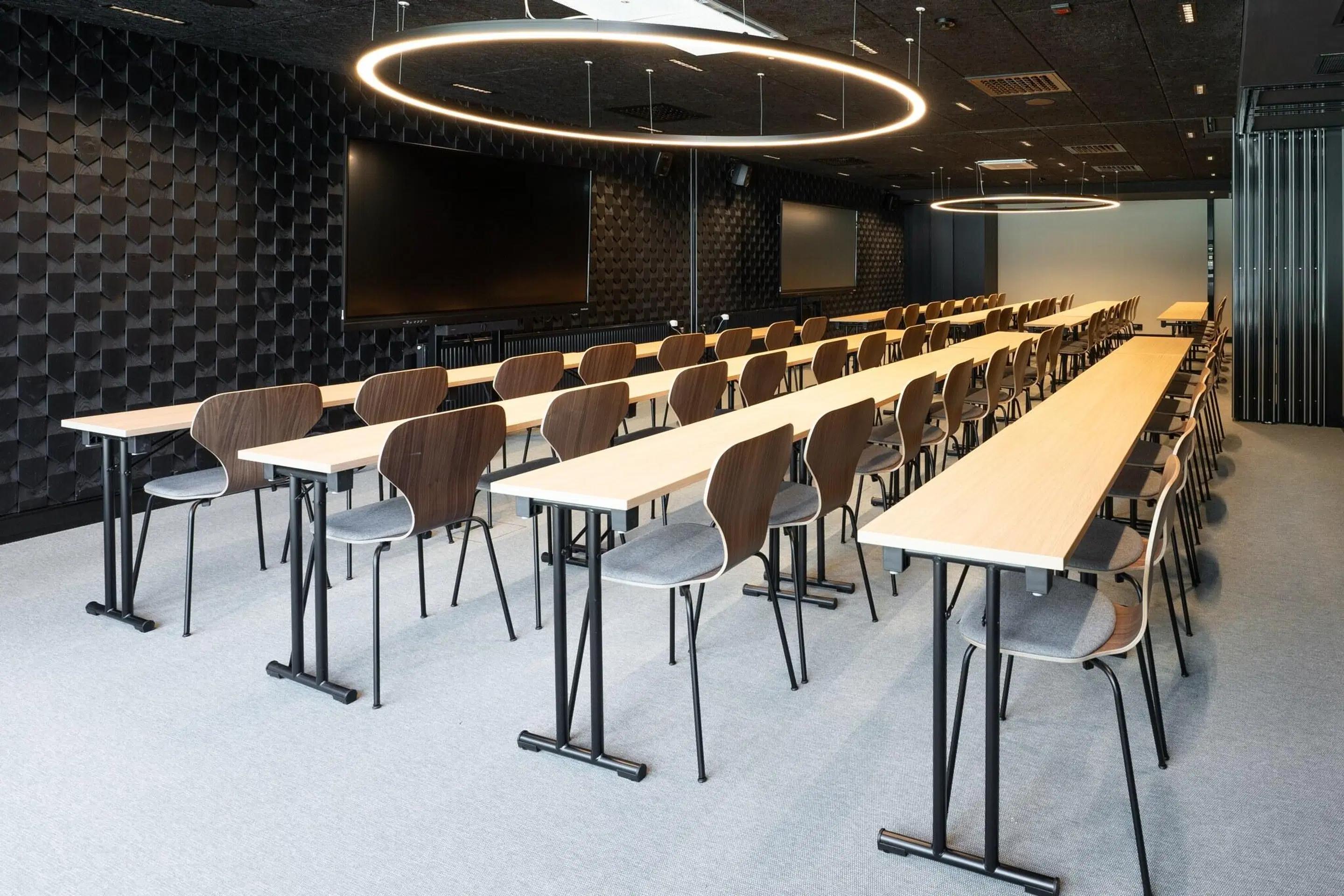 Modern auditorium with long tables and wooden-backed chairs arranged in rows. Dark acoustic panels on walls and circular ring lights hanging from ceiling.