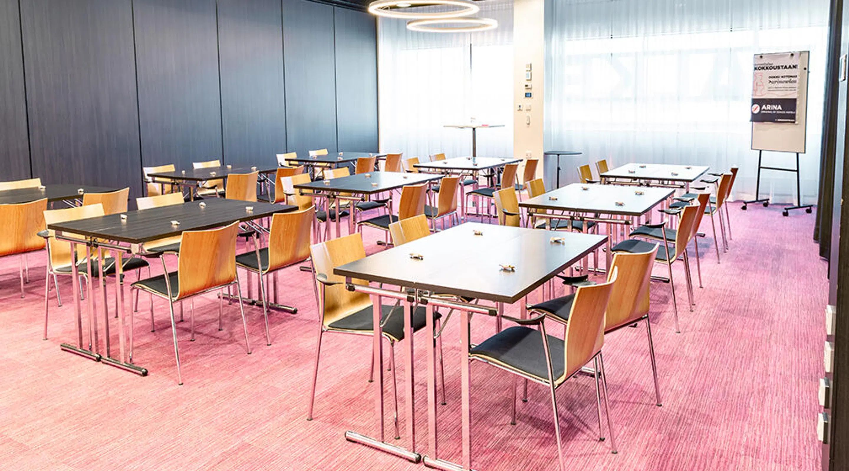 Meeting room with multiple tables and wooden chairs on pink carpeting, dark wall panels and circular ceiling lights.