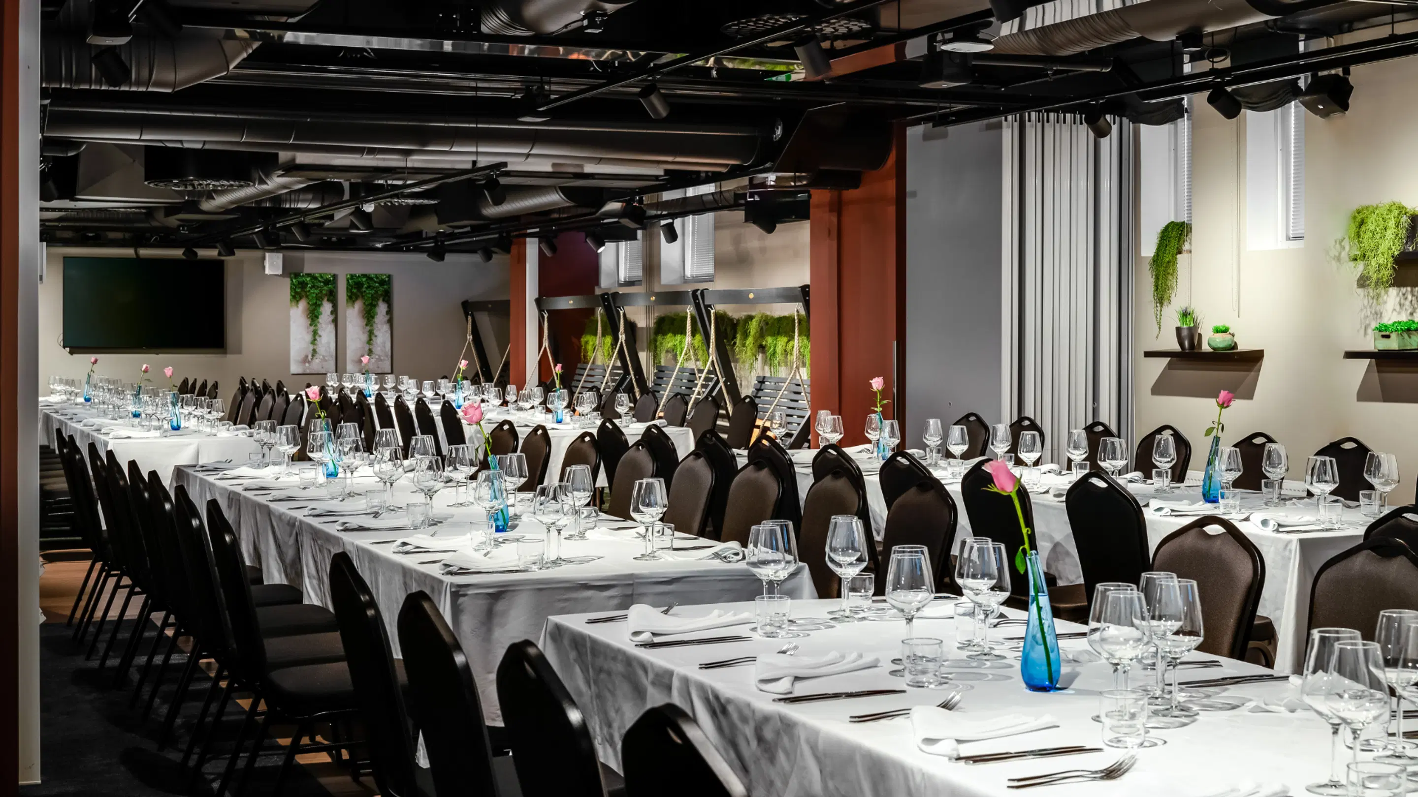Banquet room with long tables covered in white tablecloths, set with wine glasses and cutlery. The space features green plants and pink flowers in vases.
