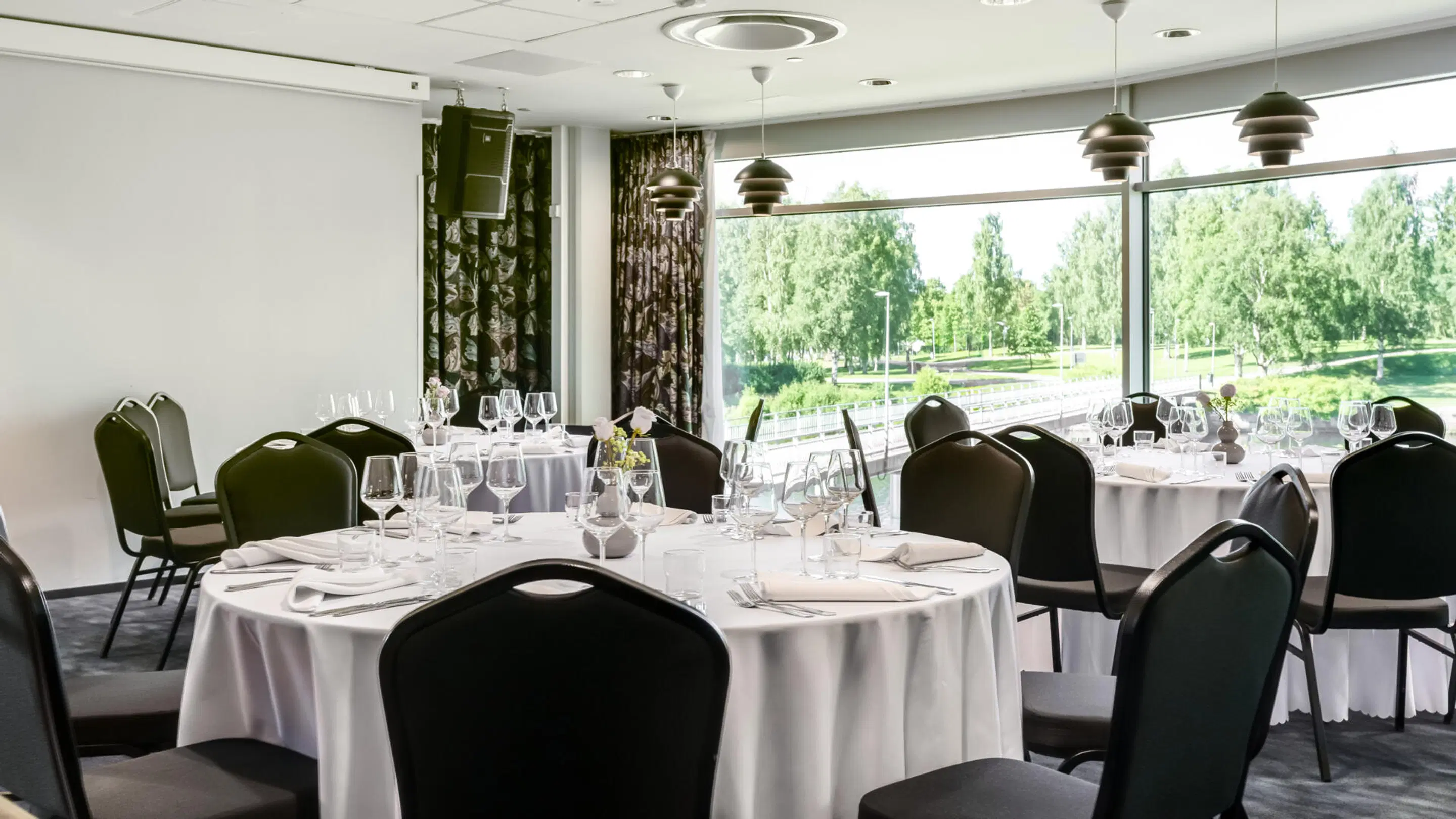 Bright banquet room with round tables, white tablecloths and black chairs. Large windows overlook a lush green park landscape.