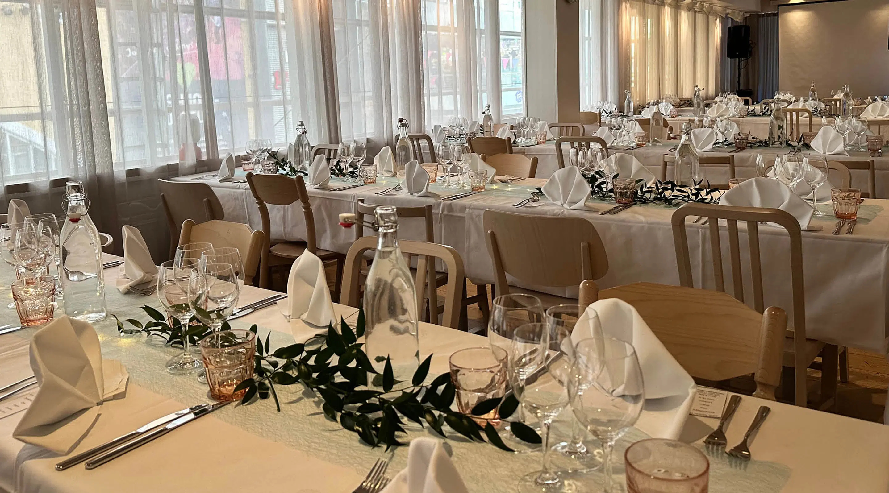 Banquet hall with multiple tables set with white tablecloths. Wooden chairs, wine glasses, napkins and green leaf decorations on tables. Large windows letting in light.