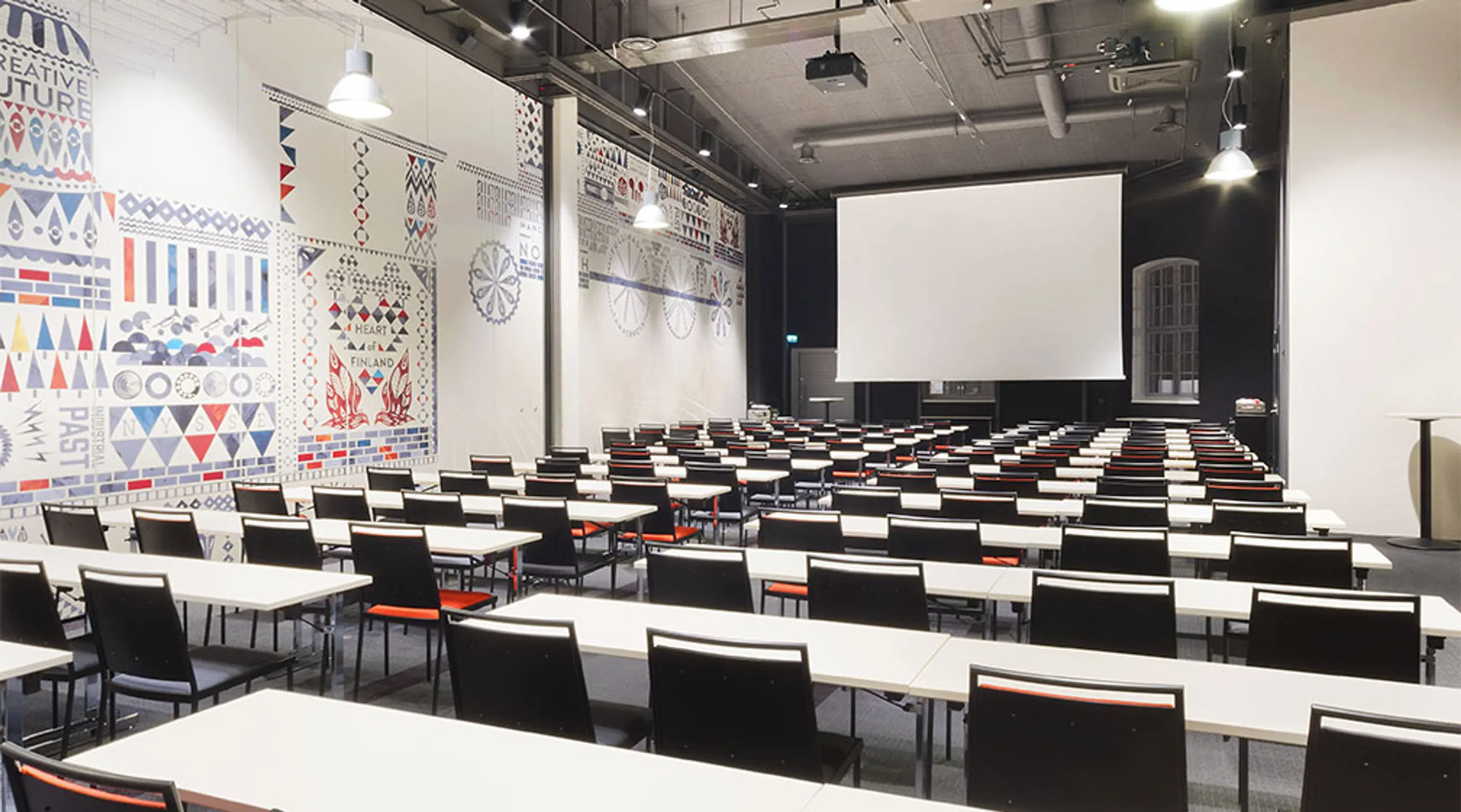 Moodne auditoorium laudade ridadega, millel on mustad toolid ja valged lauaplaadid. Seinal värvilised geomeetrilised mustrid ja ees valge projektsiooniekraan.