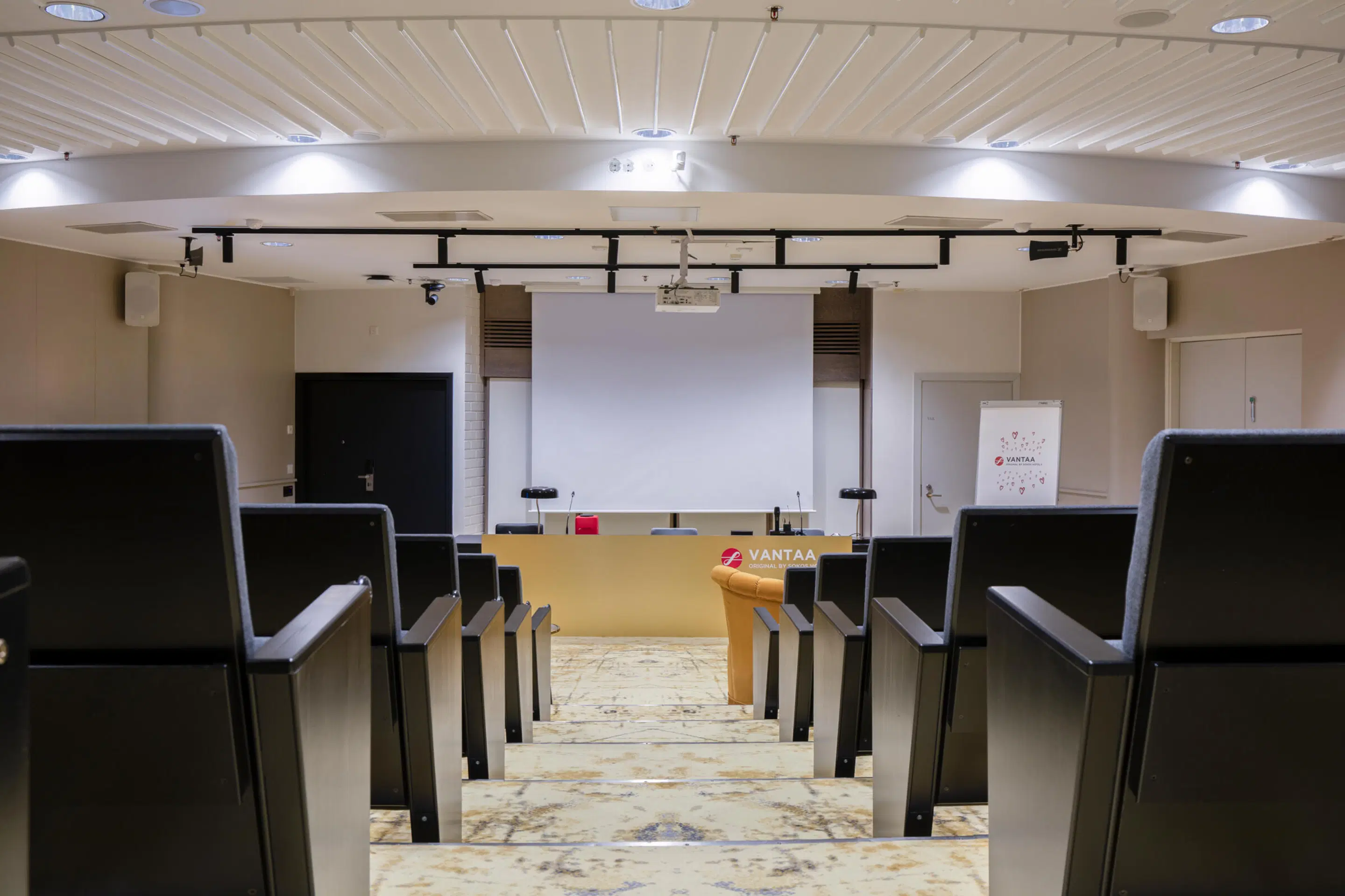 Väike auditoorium astmeliste istmetega, ees projektsiooniekraan ja esinejapult. Marmormustriline põrand ja hea laevalgustus.
