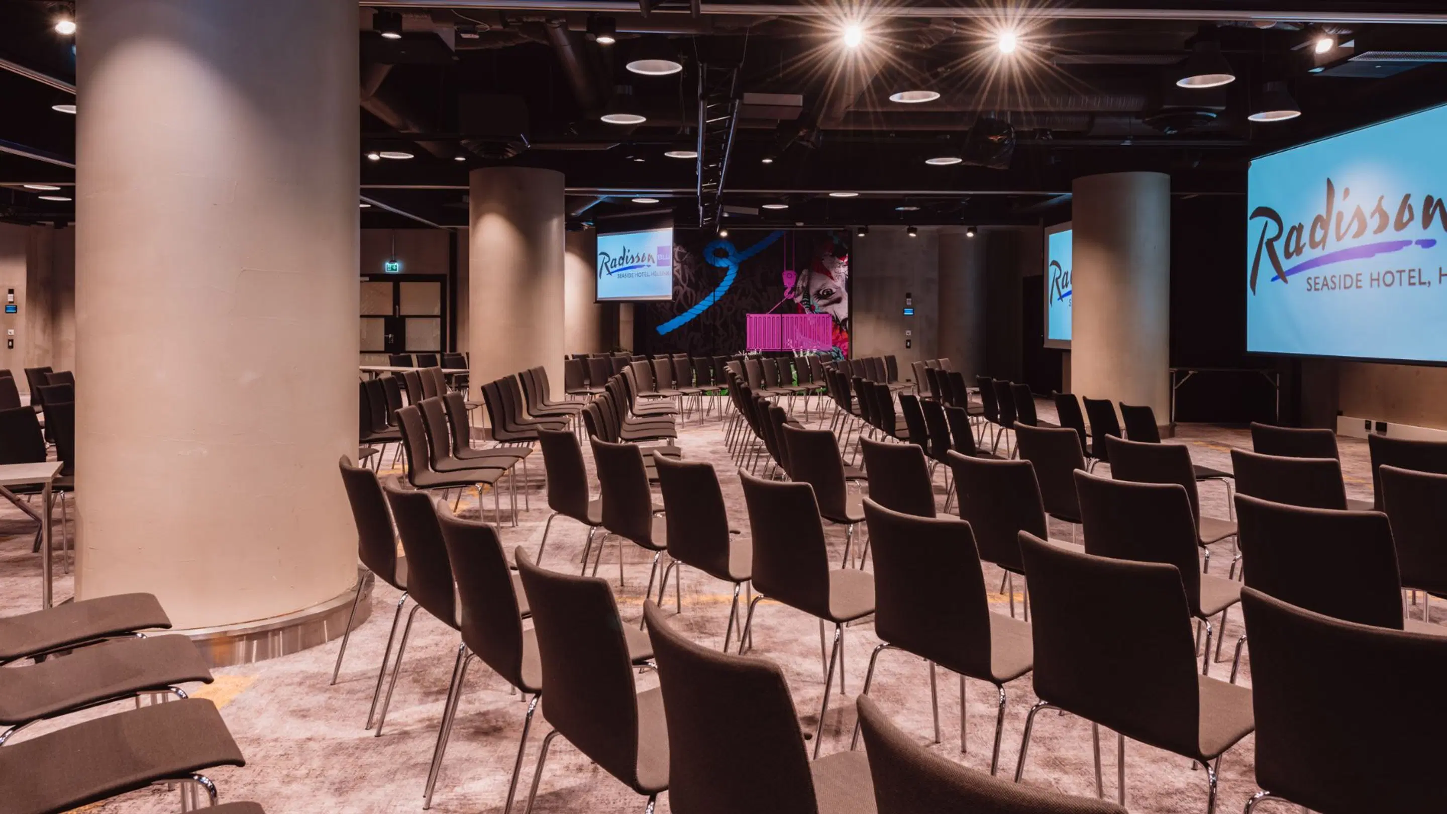 Modern auditorium in Radisson hotel with rows of chairs, three white pillars and multiple screens. Level flooring, good lighting.