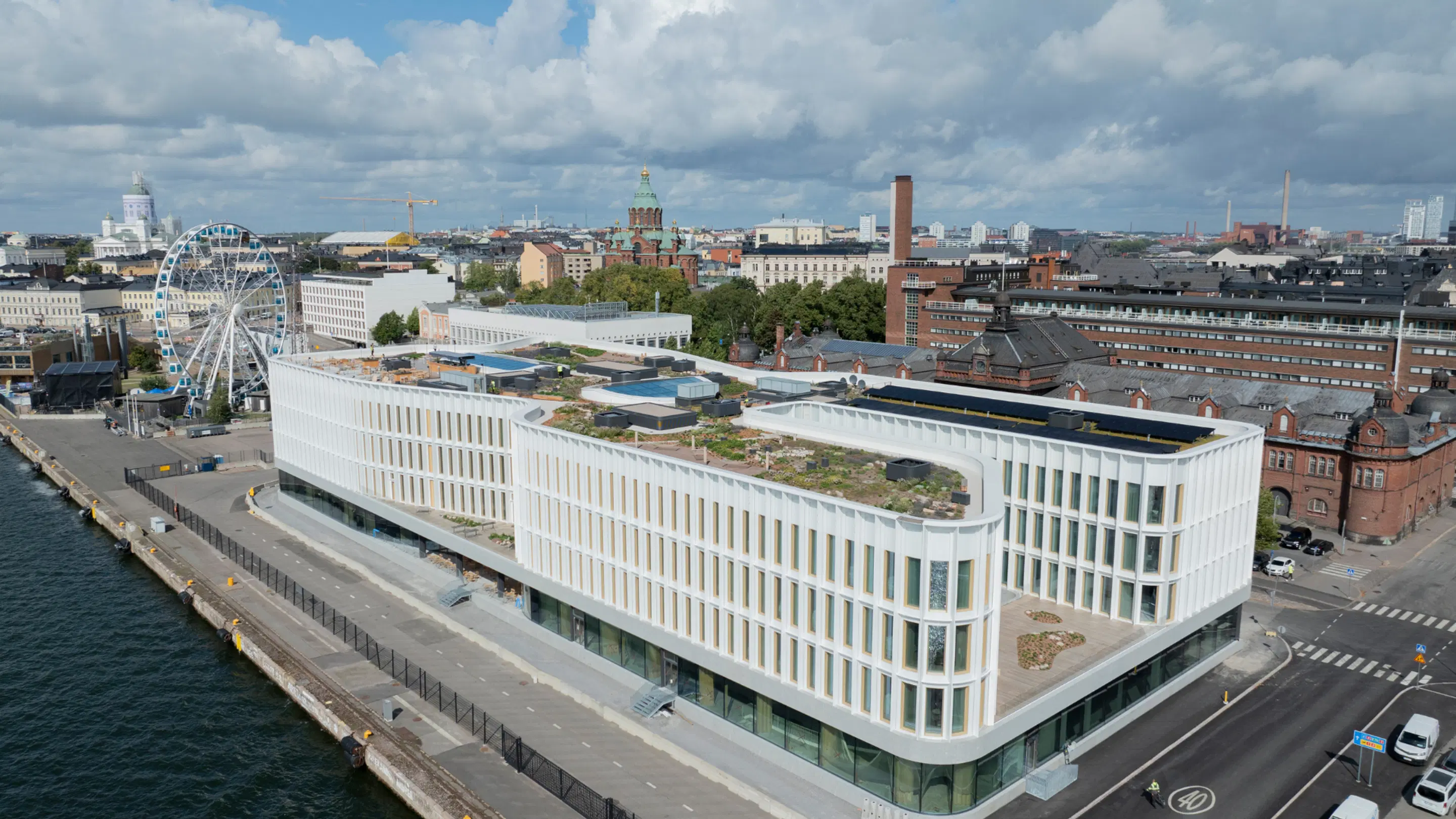 Aerial view of a modern waterfront building with green roof. Helsinki cityscape in background with ferris wheel and Helsinki Cathedral visible.