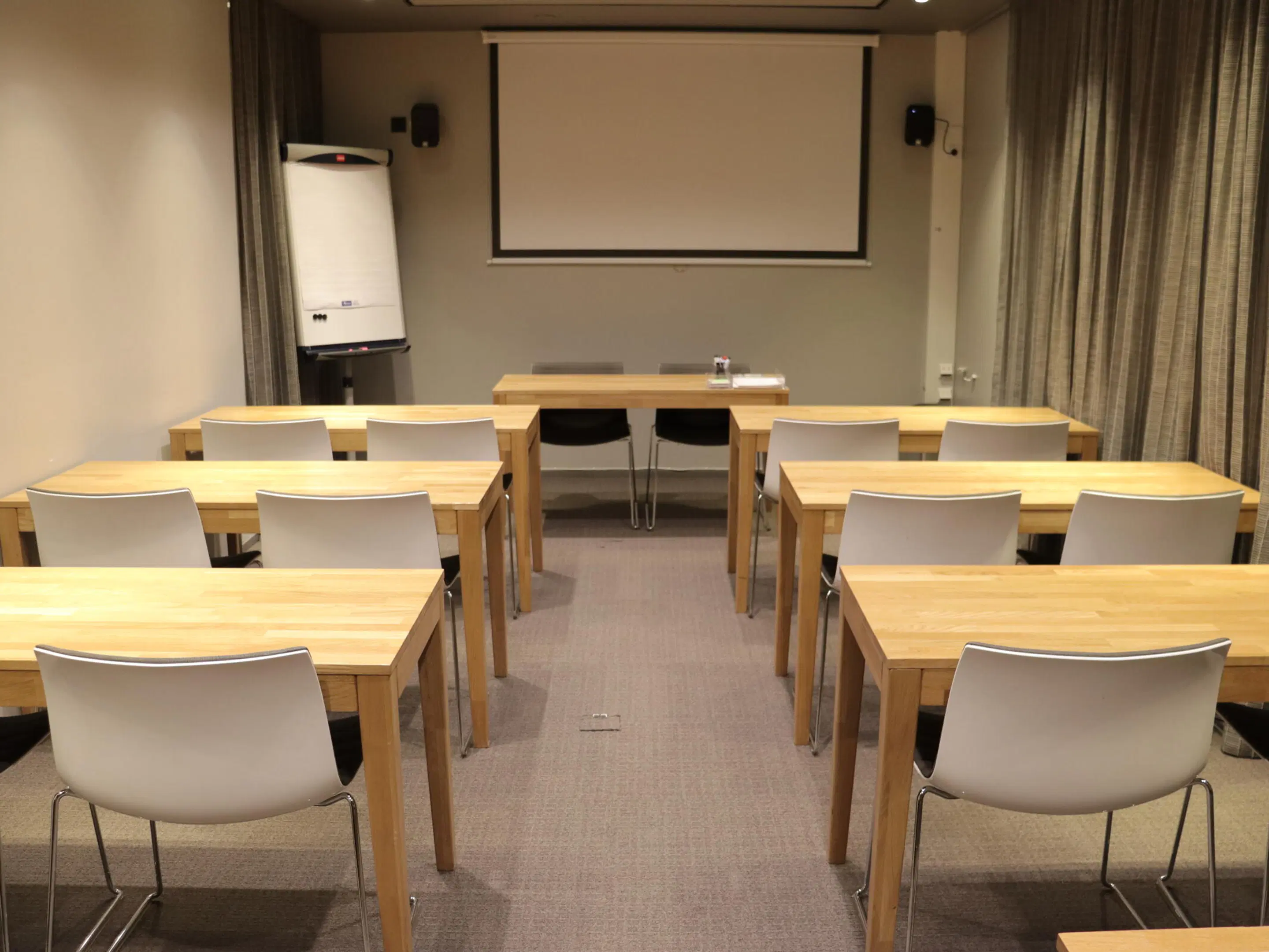 Small meeting room with wooden tables and white chairs arranged in rows facing a projection screen and flip chart. Level carpeted floor throughout.