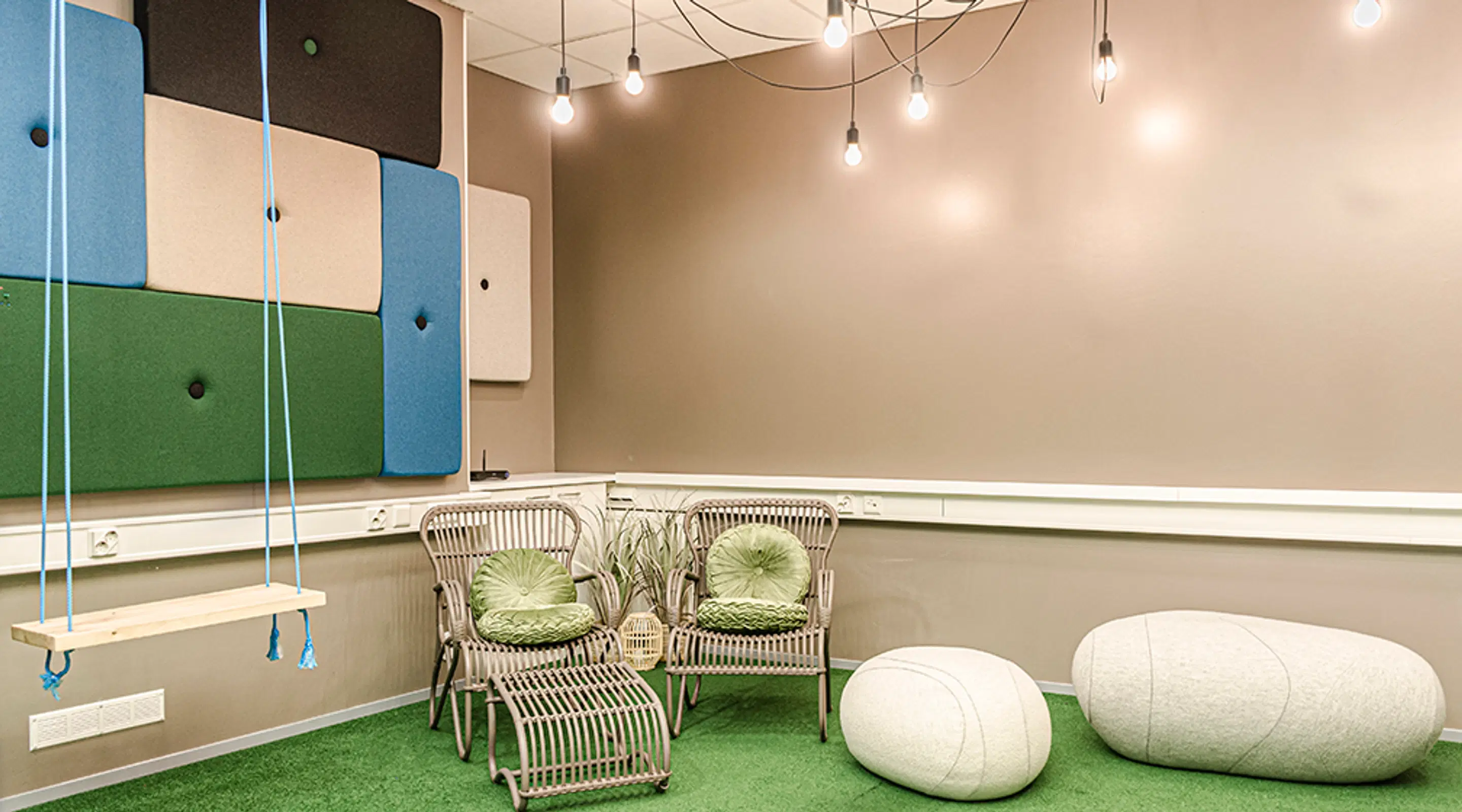 Modern lounge area with green carpet featuring swing, rattan chairs with green cushions, and white seating poufs. Colorful acoustic panels on wall.