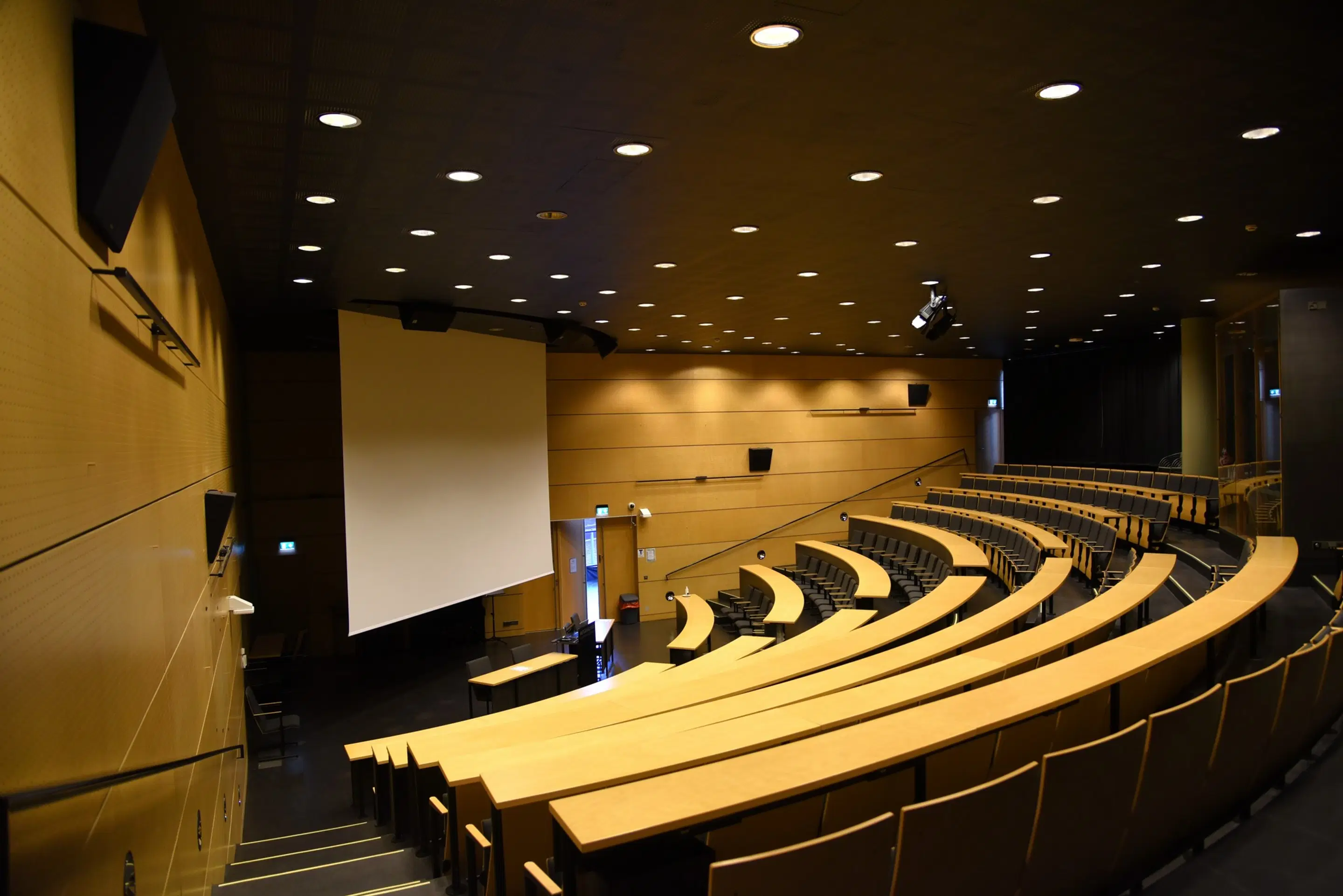 Auditoorium kaarduvate astmeliste istmeridade ja puidust laudadega. Ees on suur projektori ekraan. Treppidel on märgistatud servad ja käsipuu. Laes on süvistatud valgustid.