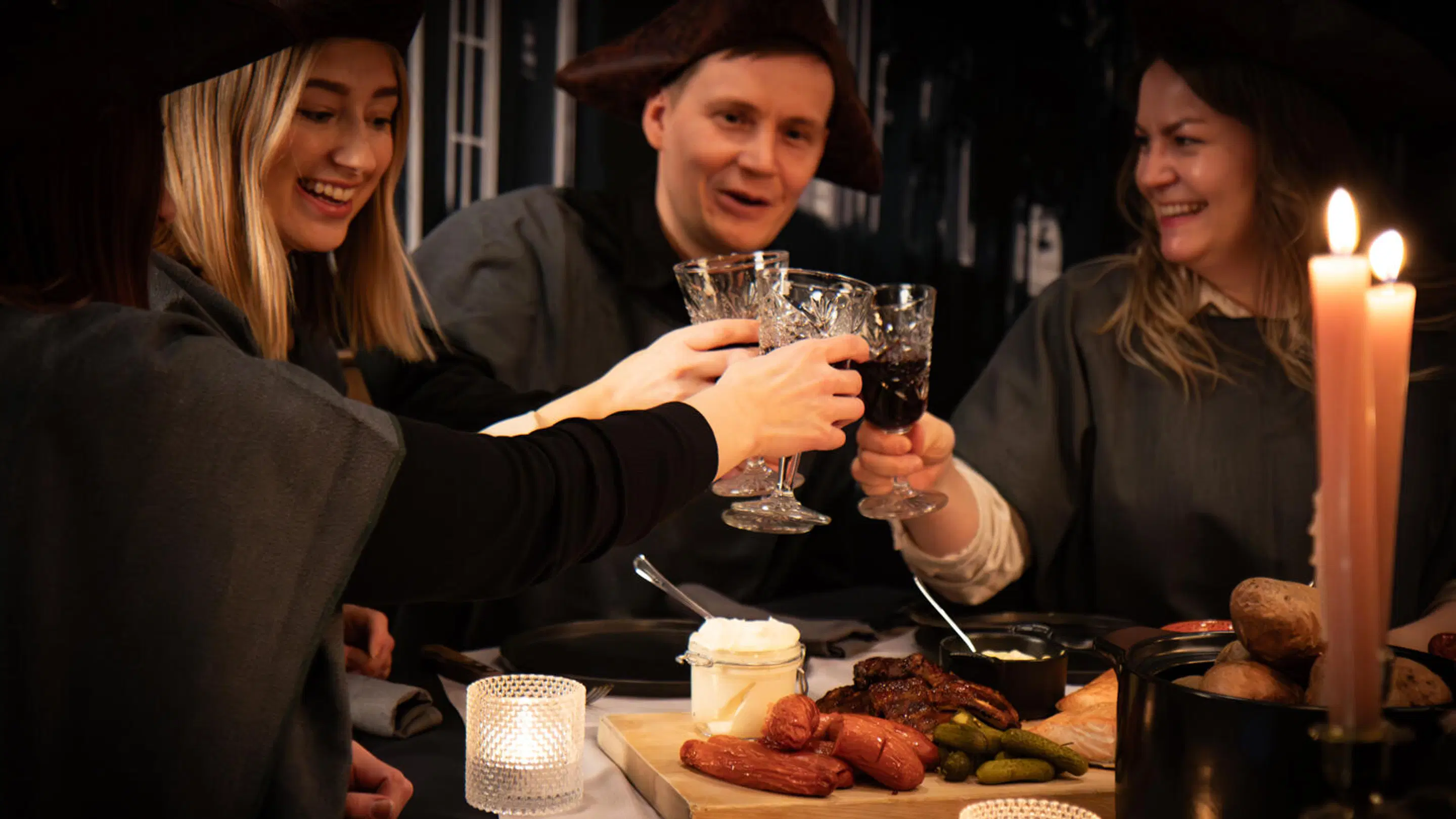 Three people in dark attire toasting crystal glasses over a platter of sausages and other foods in candlelight.