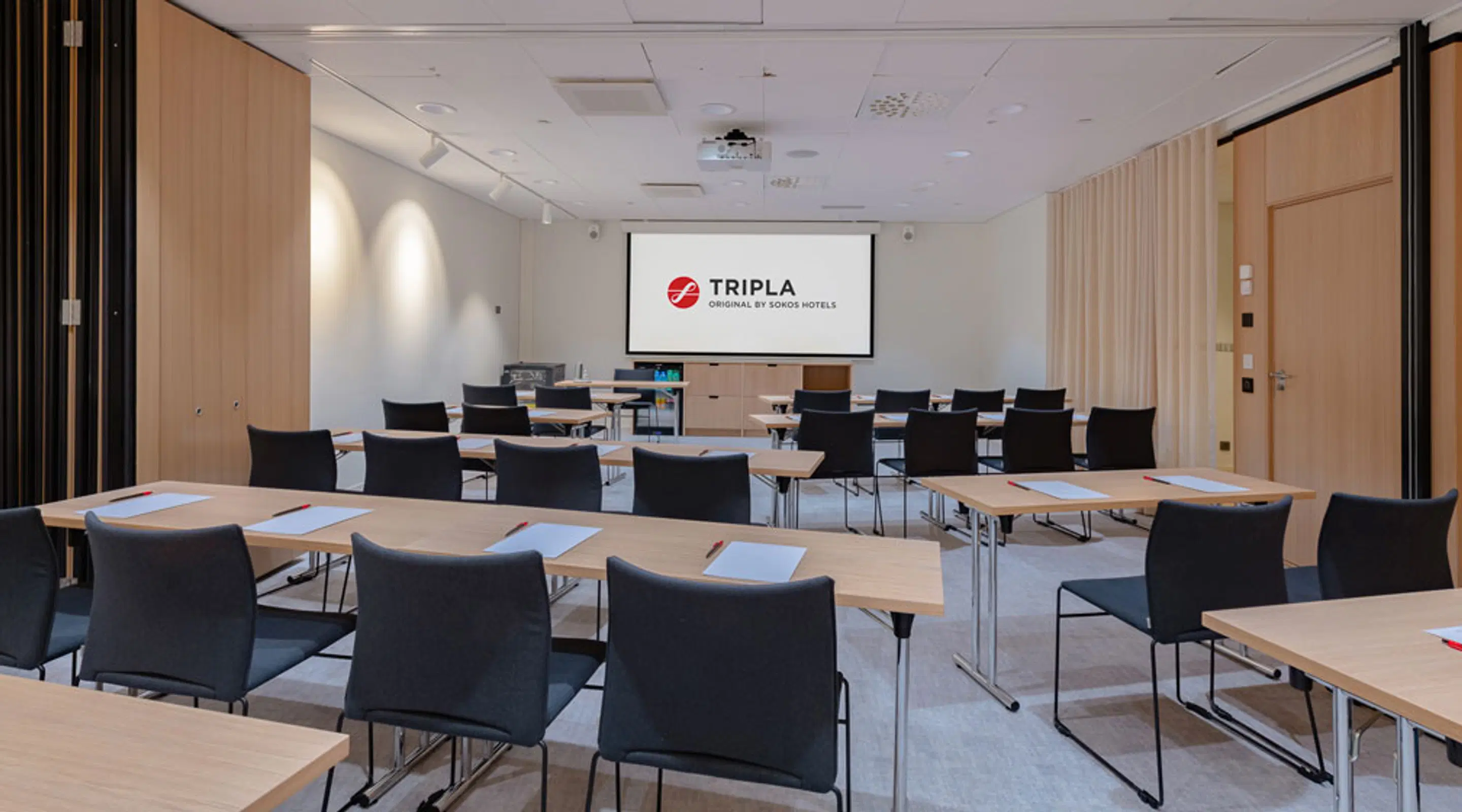 Kaasaegne koosolekuruum heledast puidust pindadega, mustade toolidega laudade ümber ja projektoriekraaniga, millel on kuvatud Tripla logo.