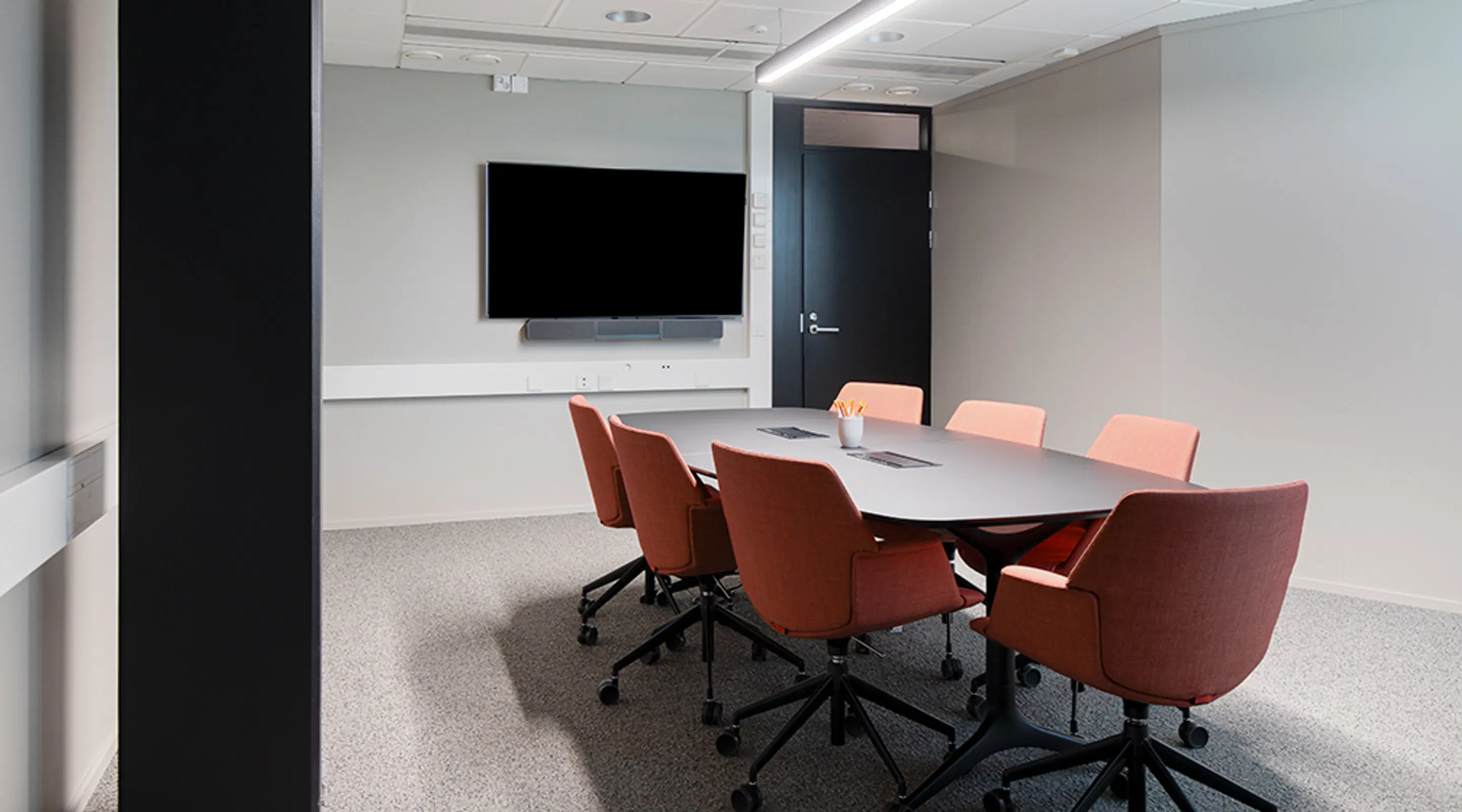 Modern conference room with oval table, terracotta-colored wheeled chairs and wall-mounted display. Room has firm floor surface and wide doorway.