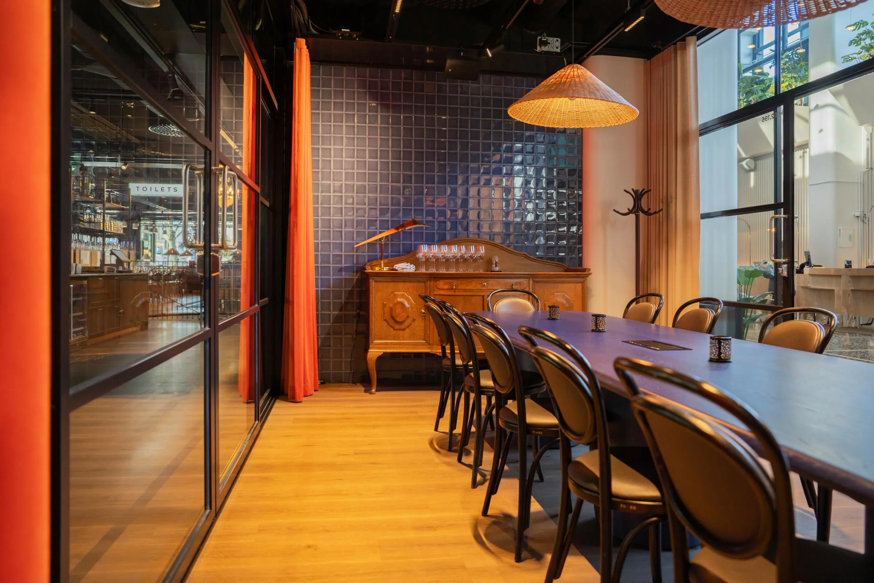 Modern meeting room with wooden floor, featuring a long dark table, decorative cabinet and wicker lamp. The space has glass walls and orange curtains.