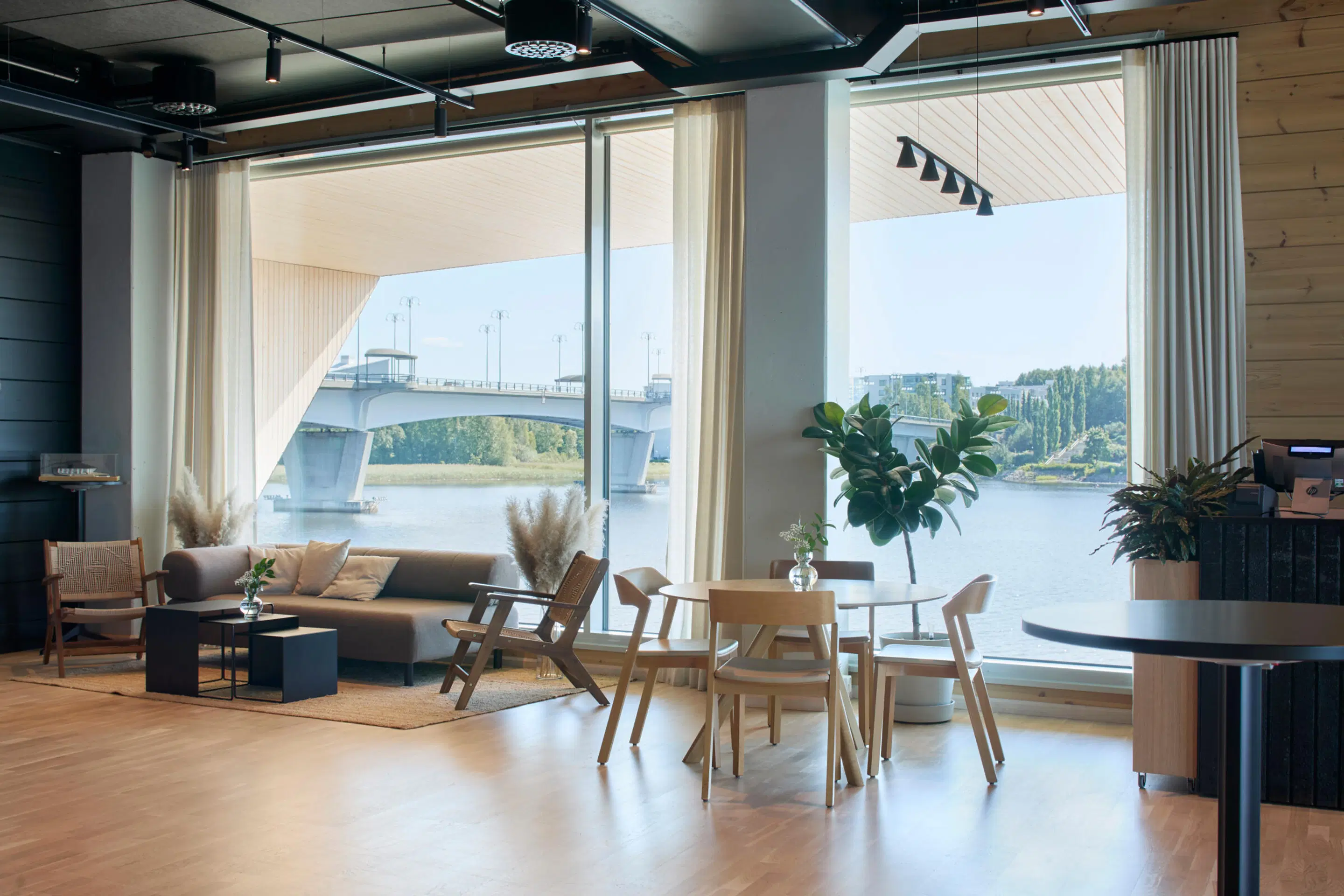 Modernt vardagsrum med trägolv, soffa, stolar och matbord. Genom stora fönster syns utsikt över en flod och bro.