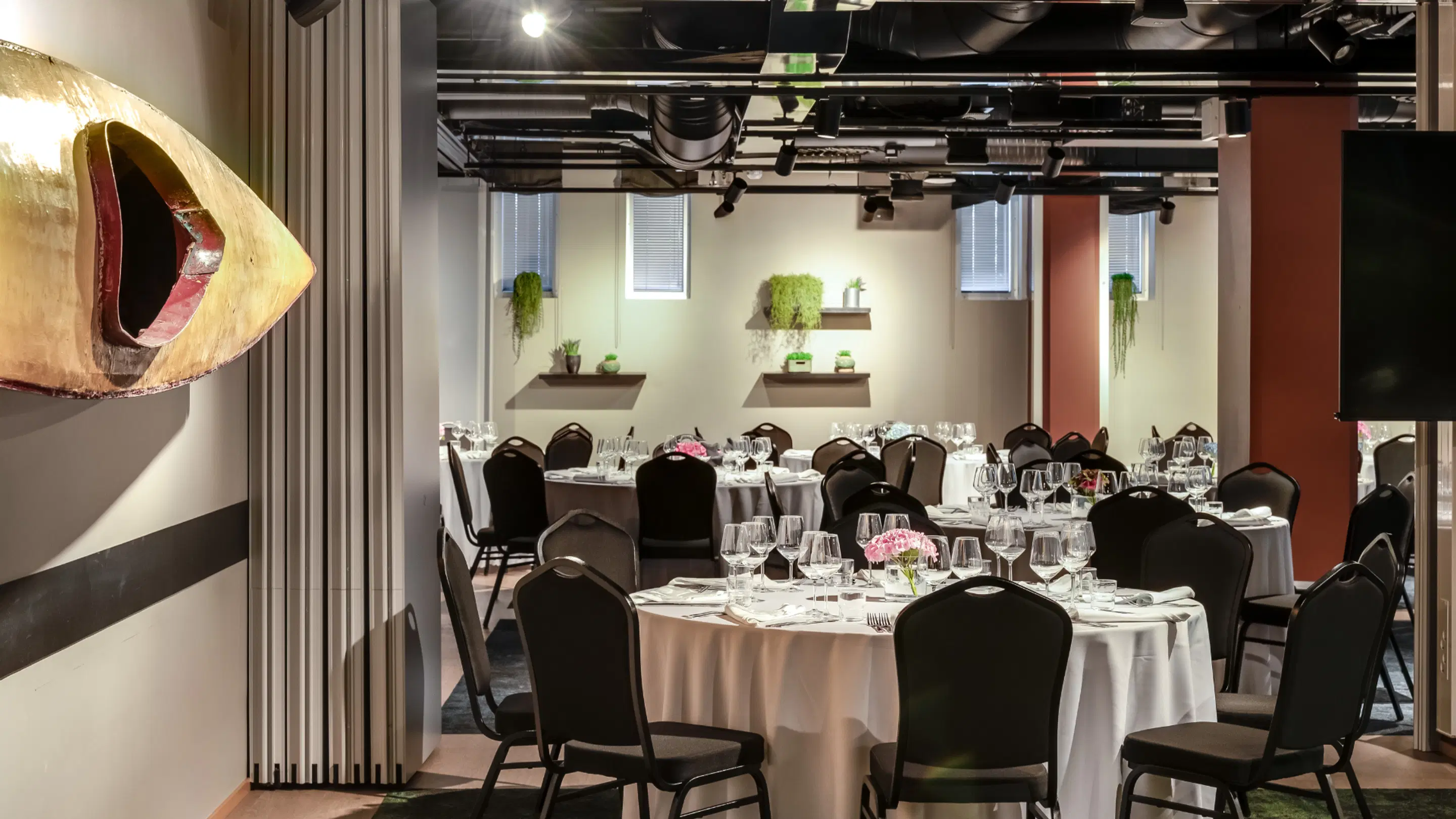 Modern banquet room with round tables covered in white tablecloths and wine glasses. A wooden art piece on the wall and green plants displayed on shelves.