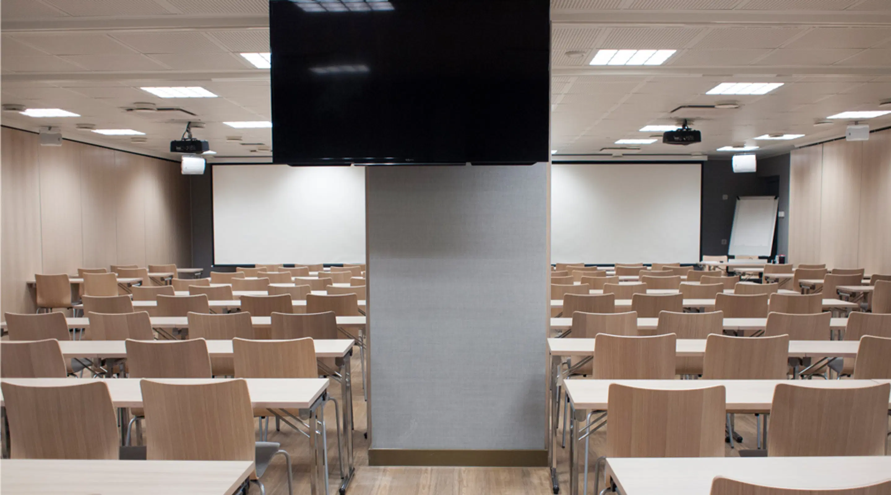 Modern auditorium med ljusa trästolar och bord, TV-skärm i mitten och vita dukar på båda sidor, rummet är väl belyst.