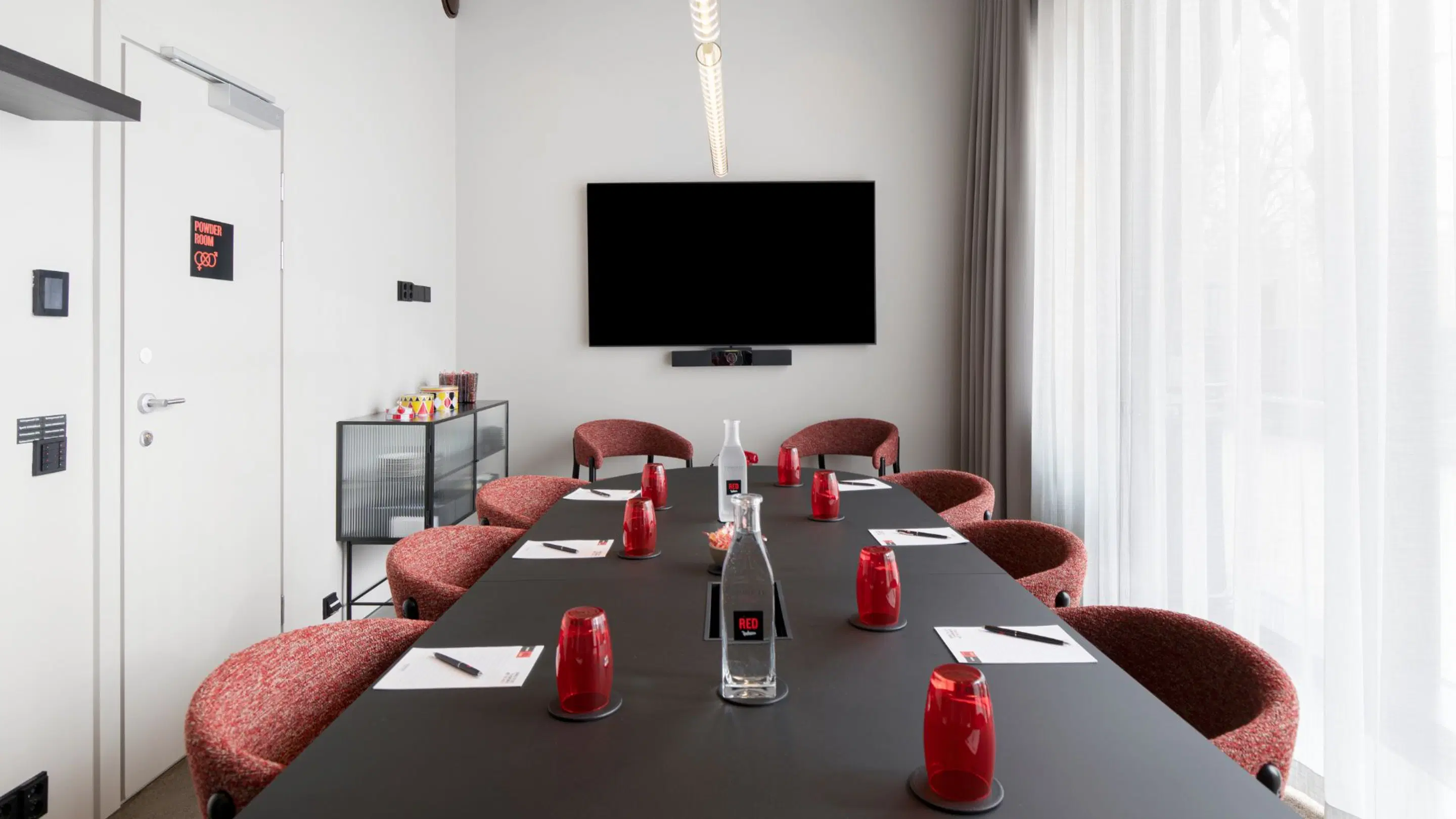 Modern meeting room with dark table, red chairs, wall-mounted display and large glass wall for natural light.