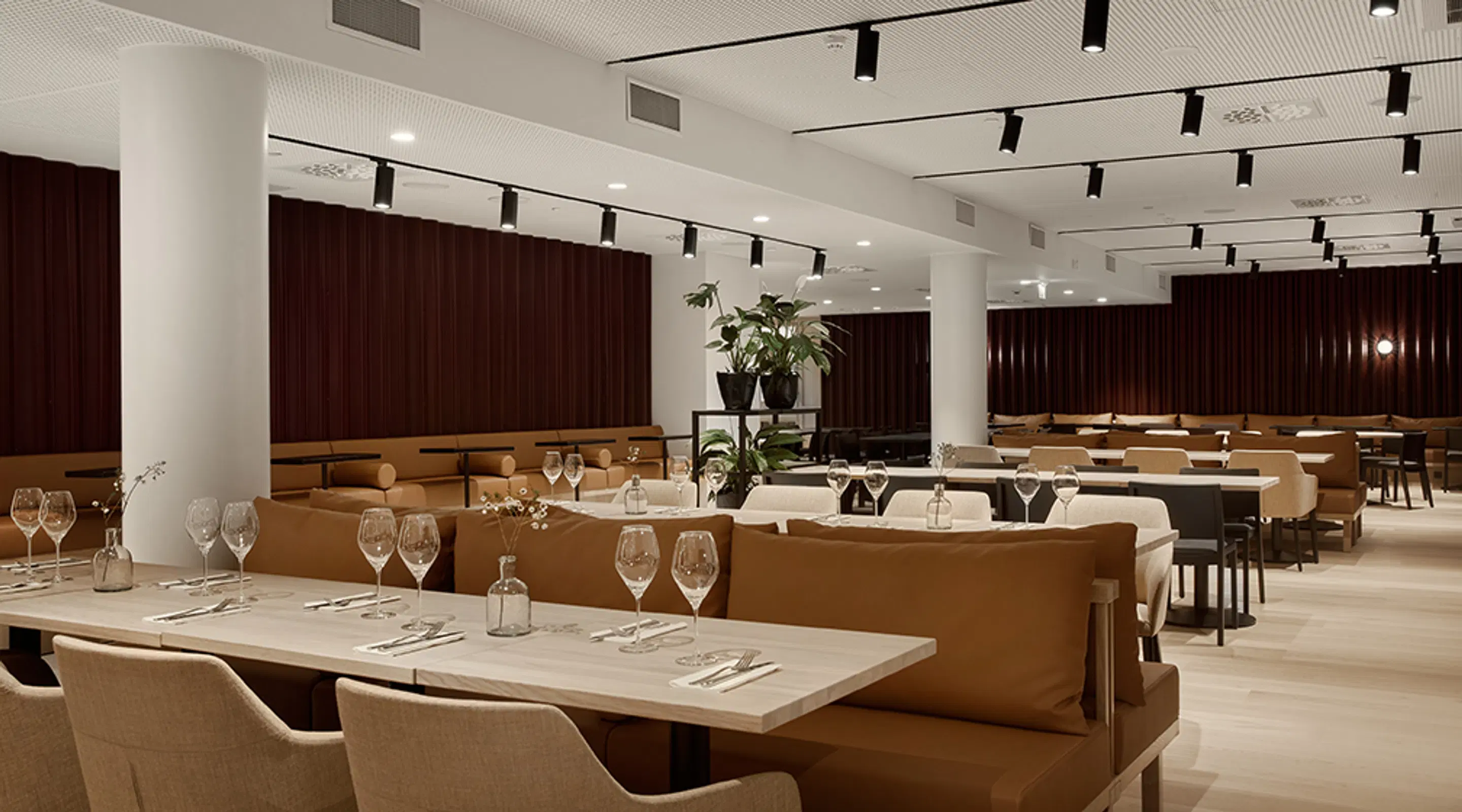 Elegant restaurant interior with light tables, brown bench seating and dark wood wall panels. Spacious area with wide pathways between tables.