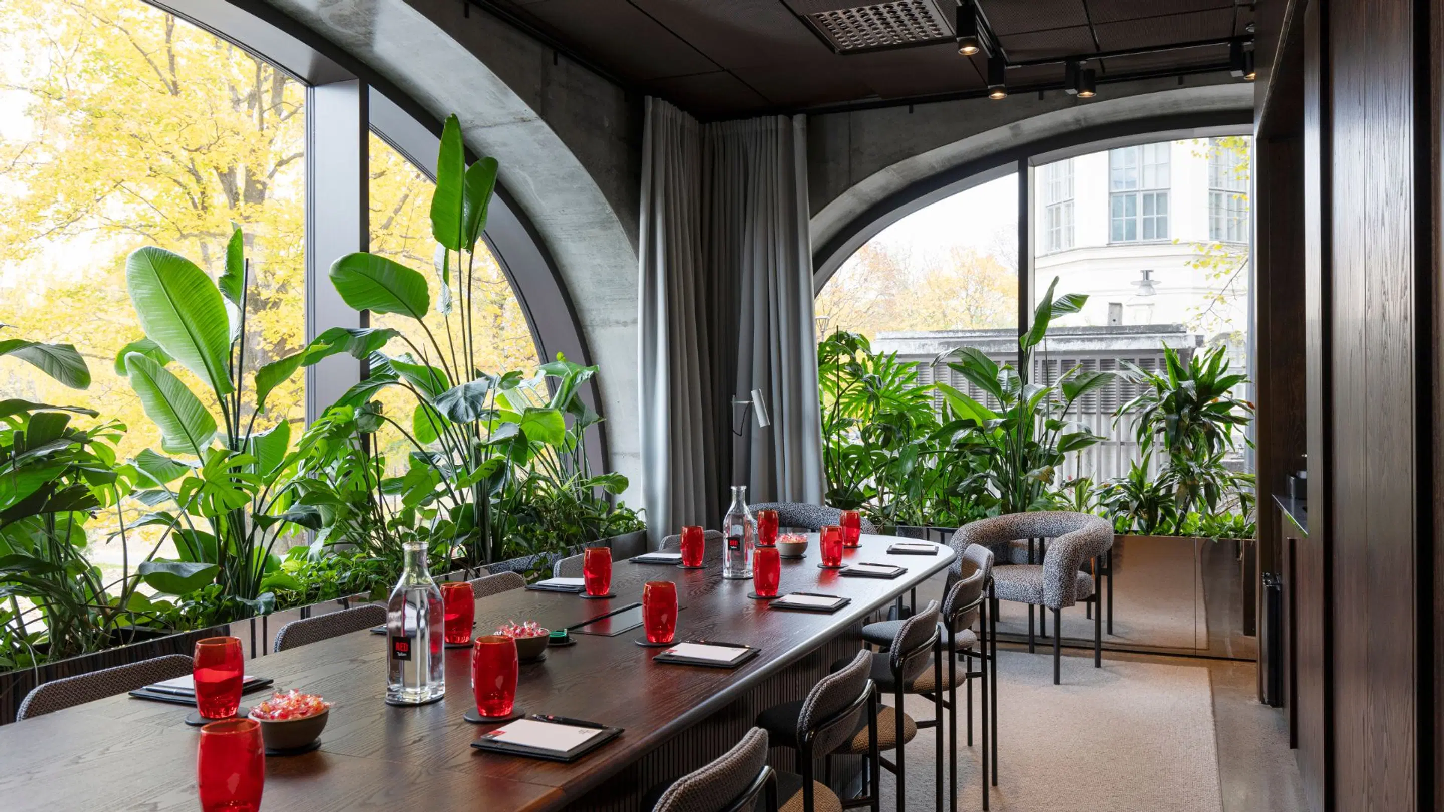 Modern meeting room with long table, red glasses and lush green plants. Arched windows showcase autumn yellow trees outside.
