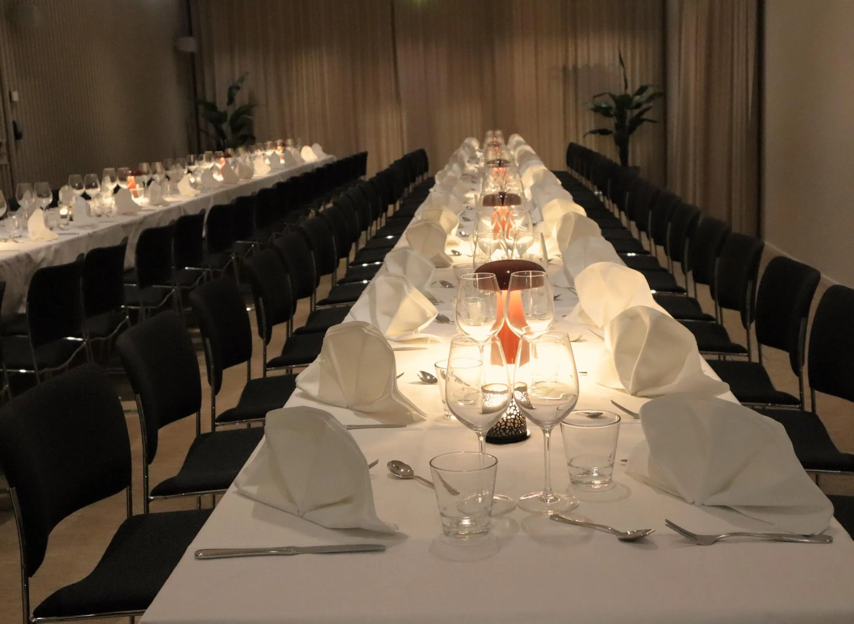 Banquet room with two long dining tables, black chairs and white tablecloths. Tables set with glassware and candles.