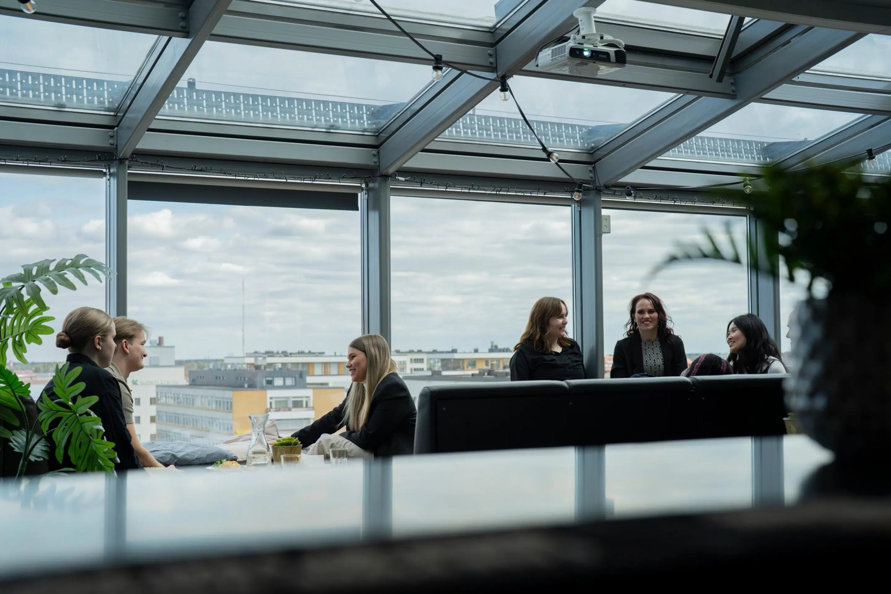A group of people conversing in a glass-roofed space with a view of the city skyline. A carafe sits on the table with plants nearby.