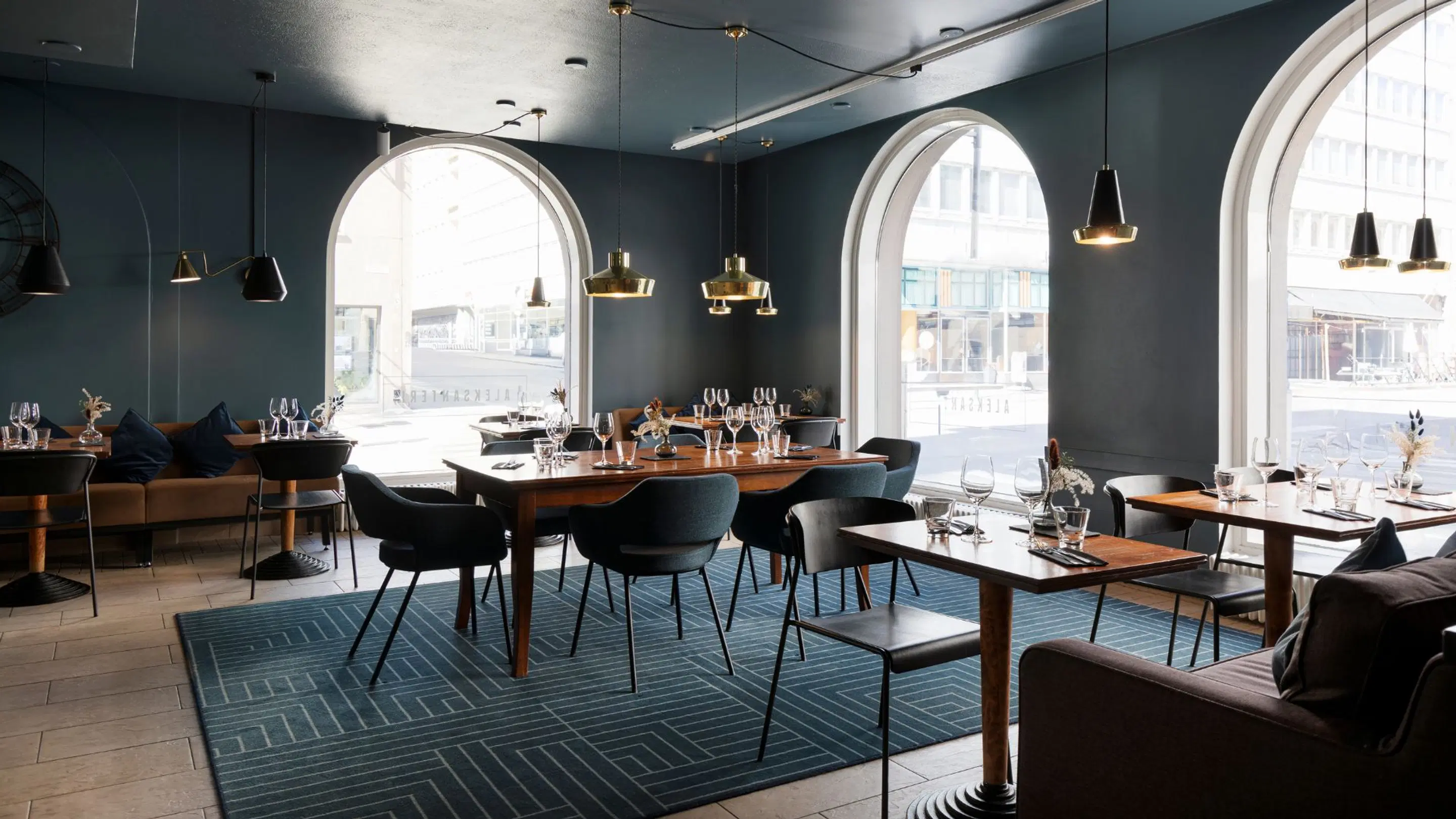 Stylish restaurant with dark grey walls and large arched windows. Wooden tables, black chairs, and elegant pendant lights create an atmospheric dining space.
