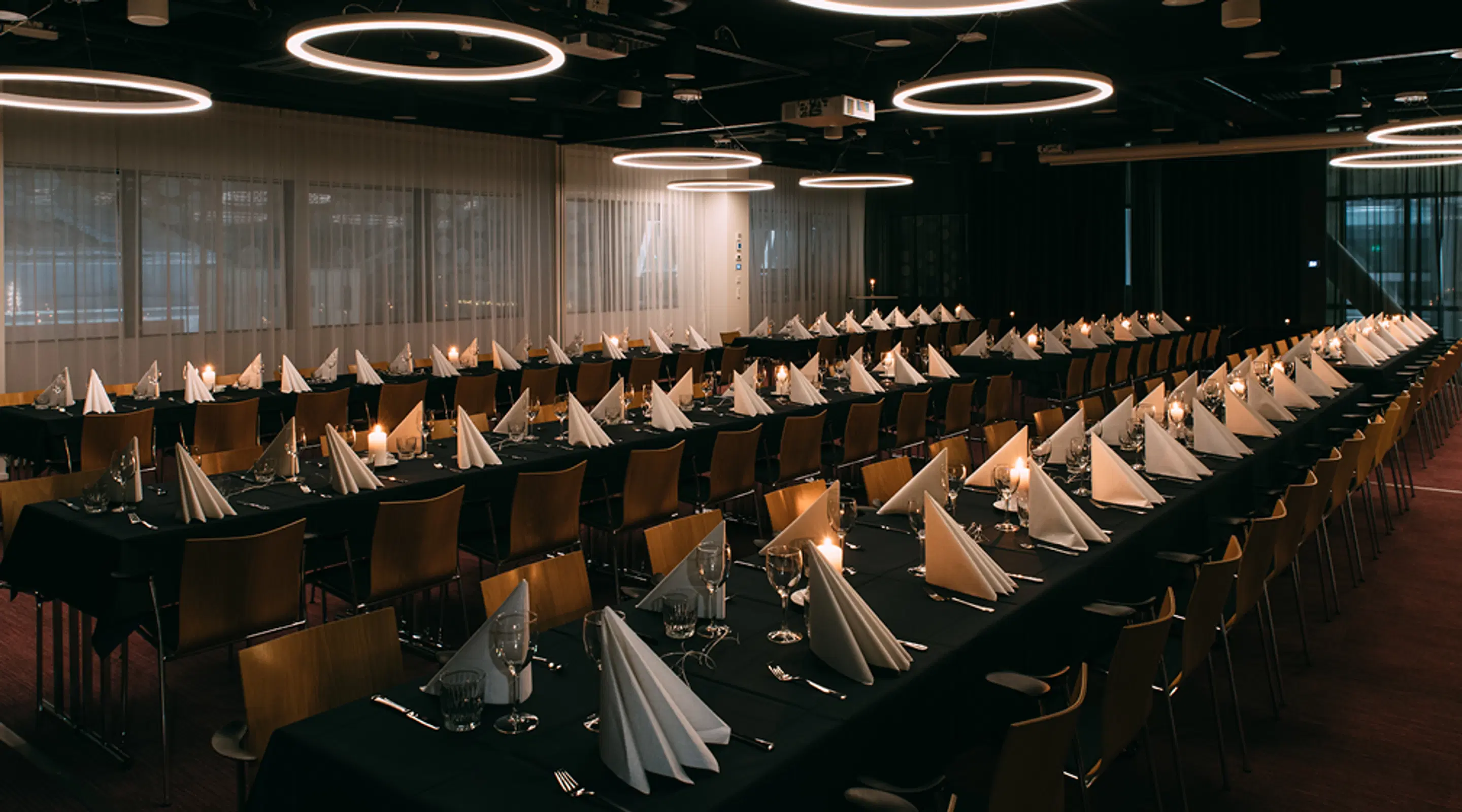 Banquet hall with long tables covered in black tablecloths, folded white napkins and place settings. Ceiling lit with circular ring lights.