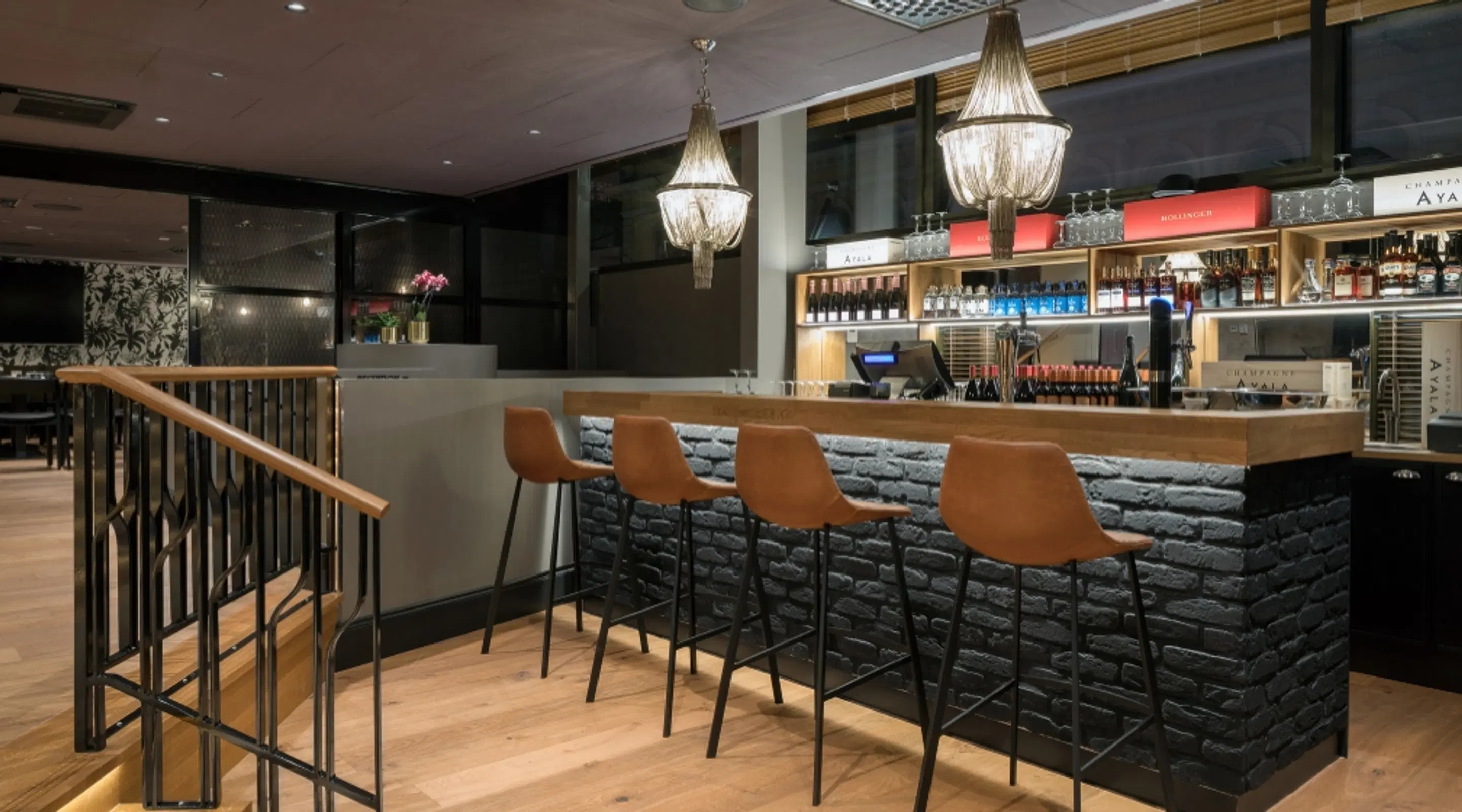 Modern bar med svart tegeldisk, orangebruna barstolar, kristallkronor och trägolv.