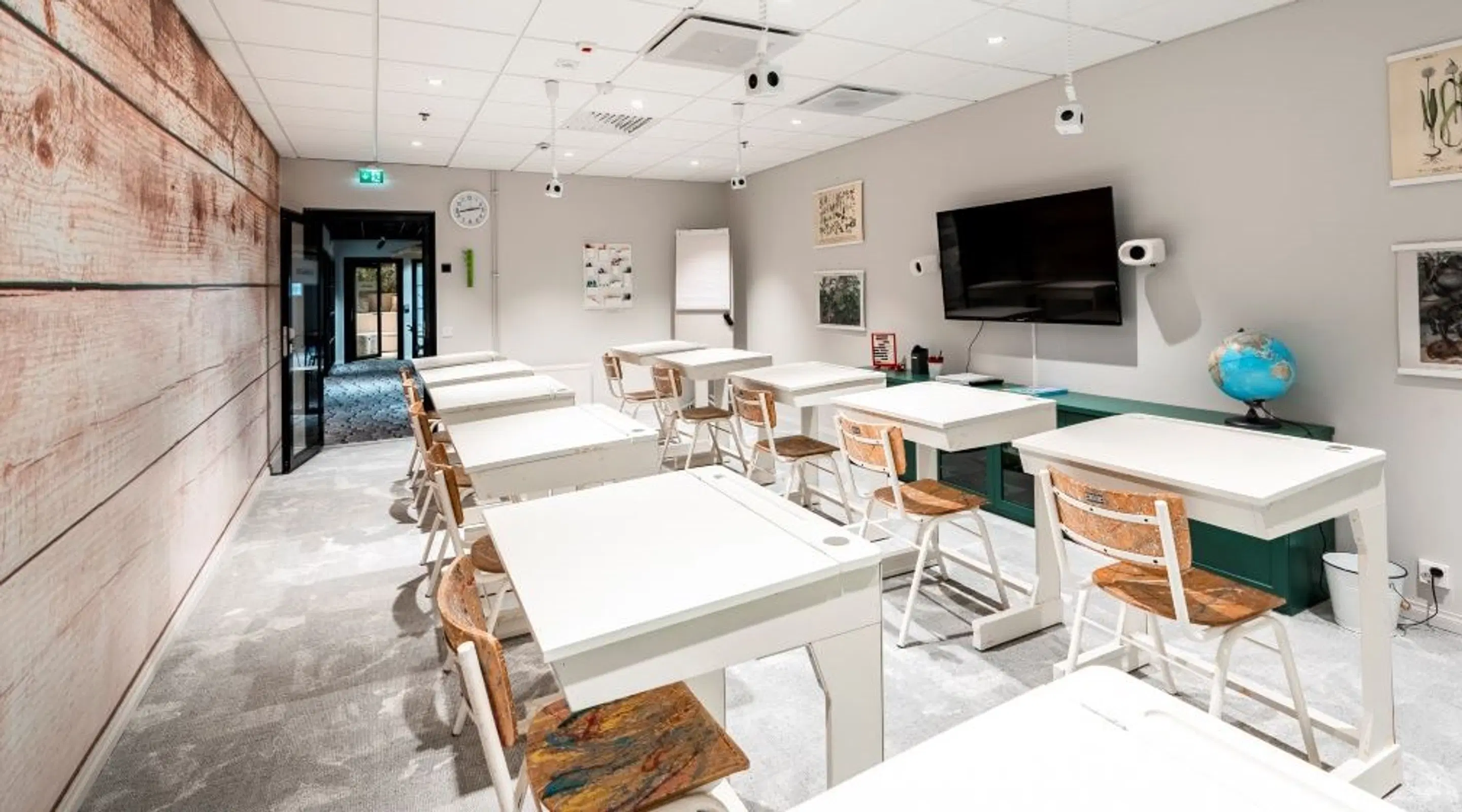 Modern classroom with white tables and wooden chairs. Wall-mounted TV, maps and botanical prints. Grey carpet flooring and wooden wall paneling.