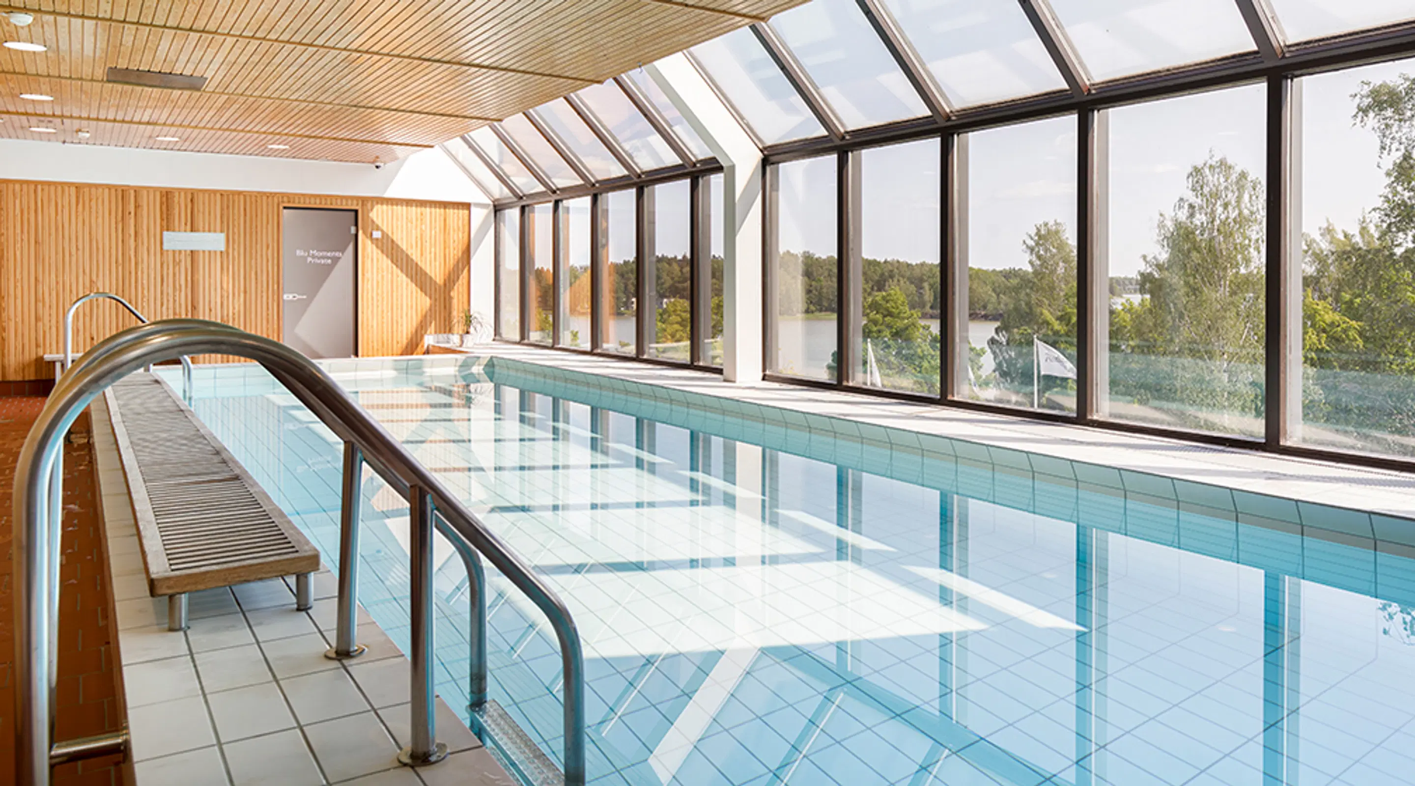 Indoor swimming pool with wooden ceiling panels and large windows showing a lake view. Metal railing and bench along the pool edge.