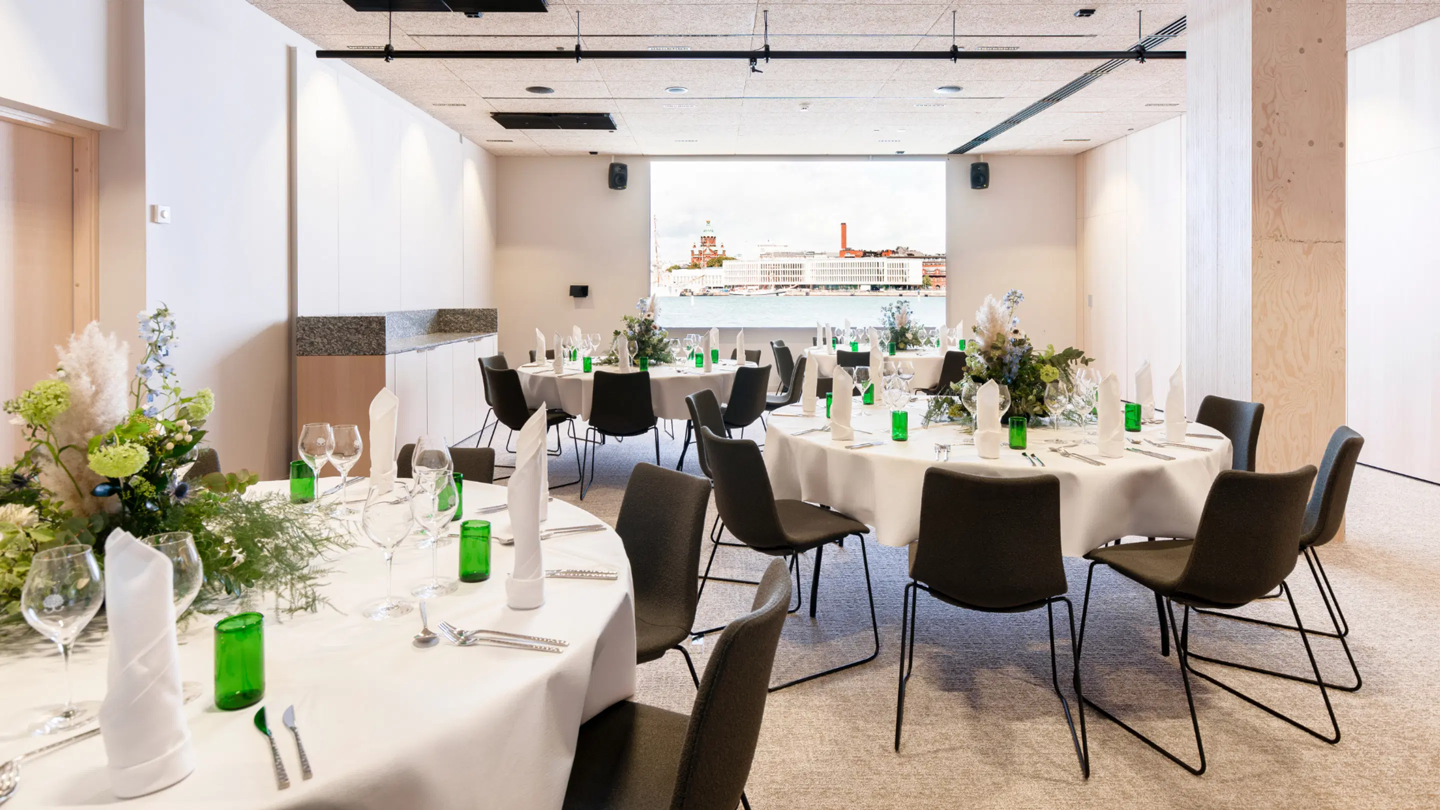 Bright event space with round tables covered in white tablecloths and black chairs. Large window showing cityscape view in background.