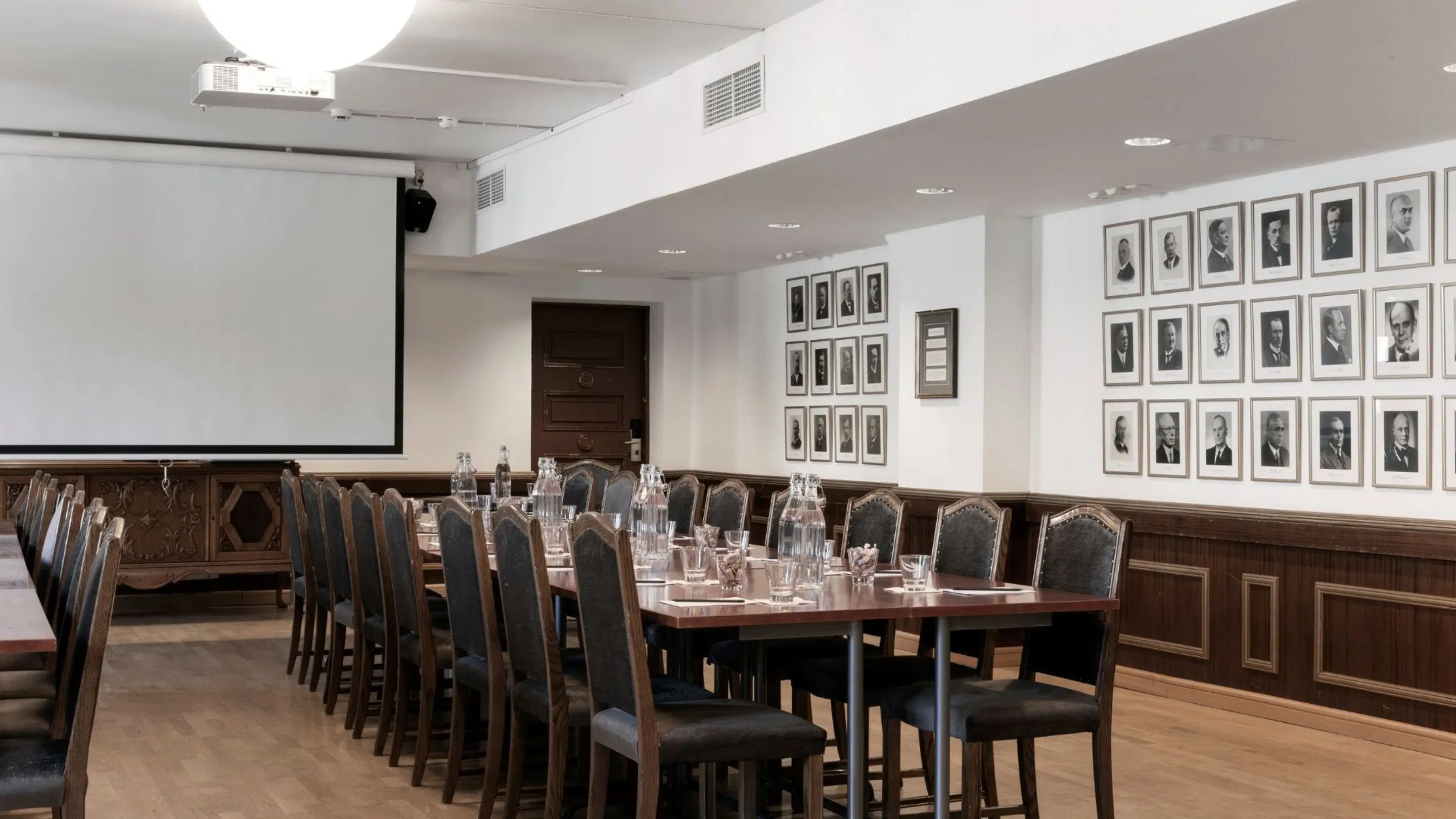 Conference room with a long table and chairs, projector, screen, and portrait photographs on walls. Wooden floor and good lighting.