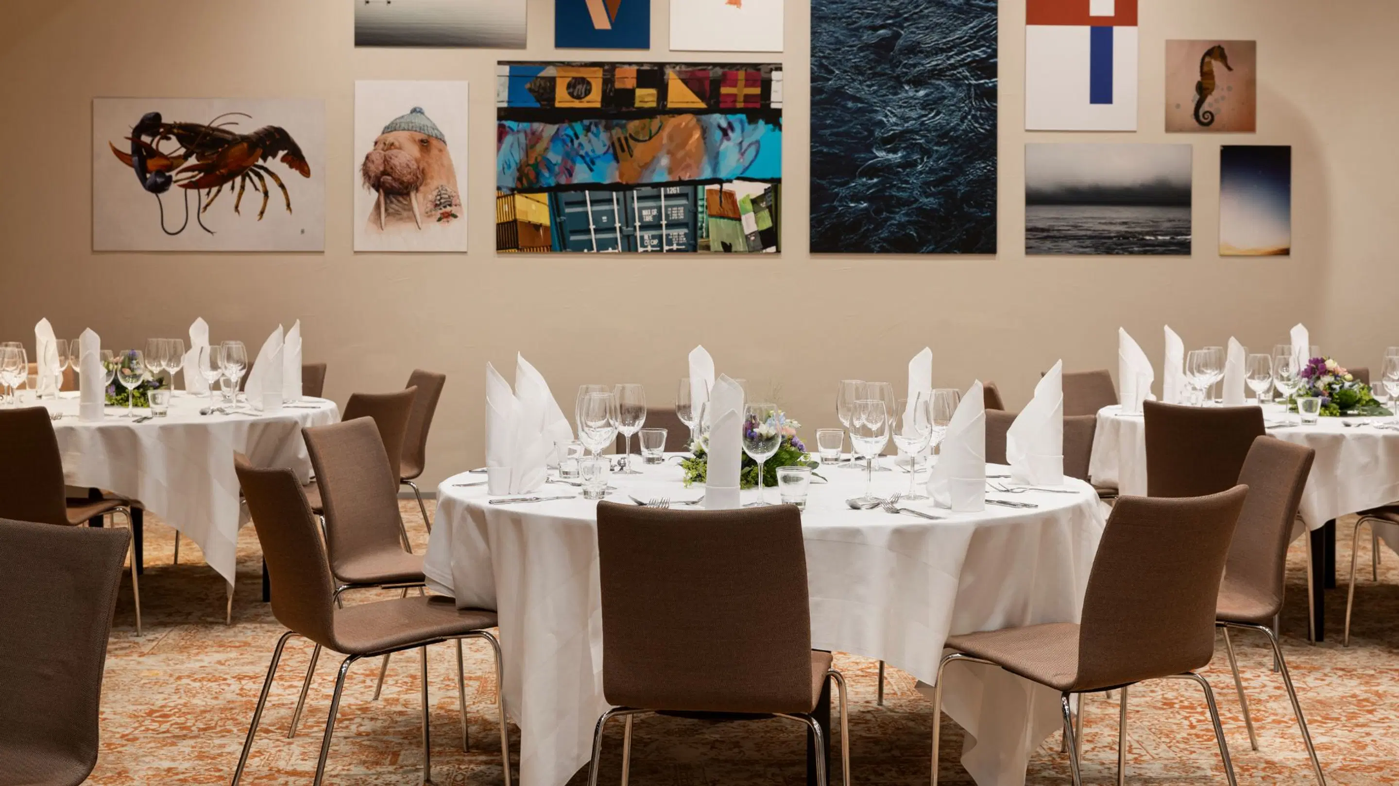 Restaurant dining room with round tables covered in white tablecloths and formal place settings. Artwork on walls, carpeted floor.