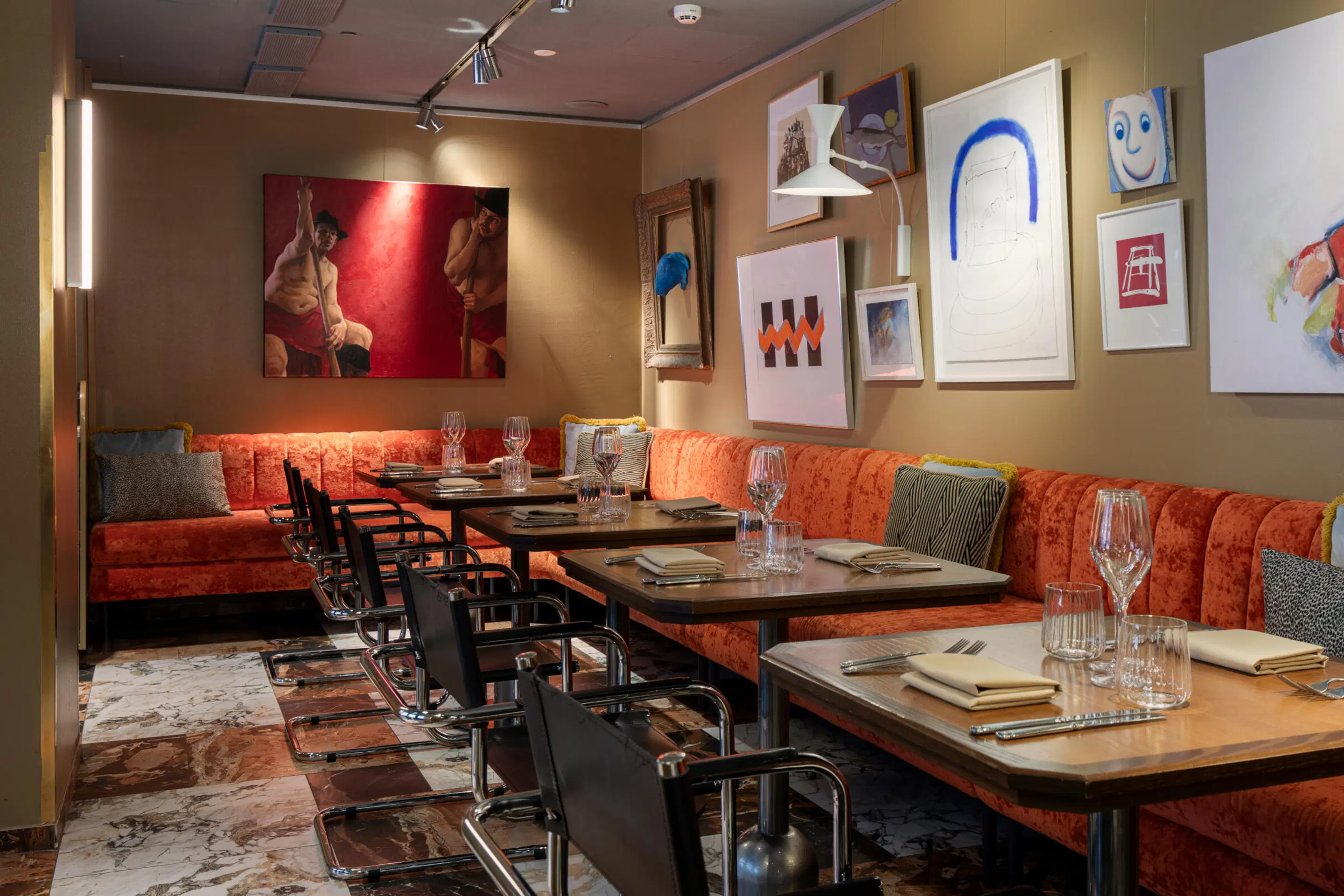 Restauranginredning med orange soffa, mörka bord och stolar. Färgglad konst på väggarna över marmorgolv.